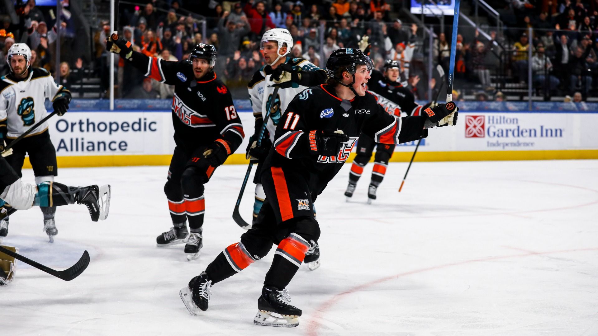 Hockey players in KC Mavericks black and orange uniforms celebrating a goal on the ice as the opposing team in white and teal looks on, with fans cheering in the background.