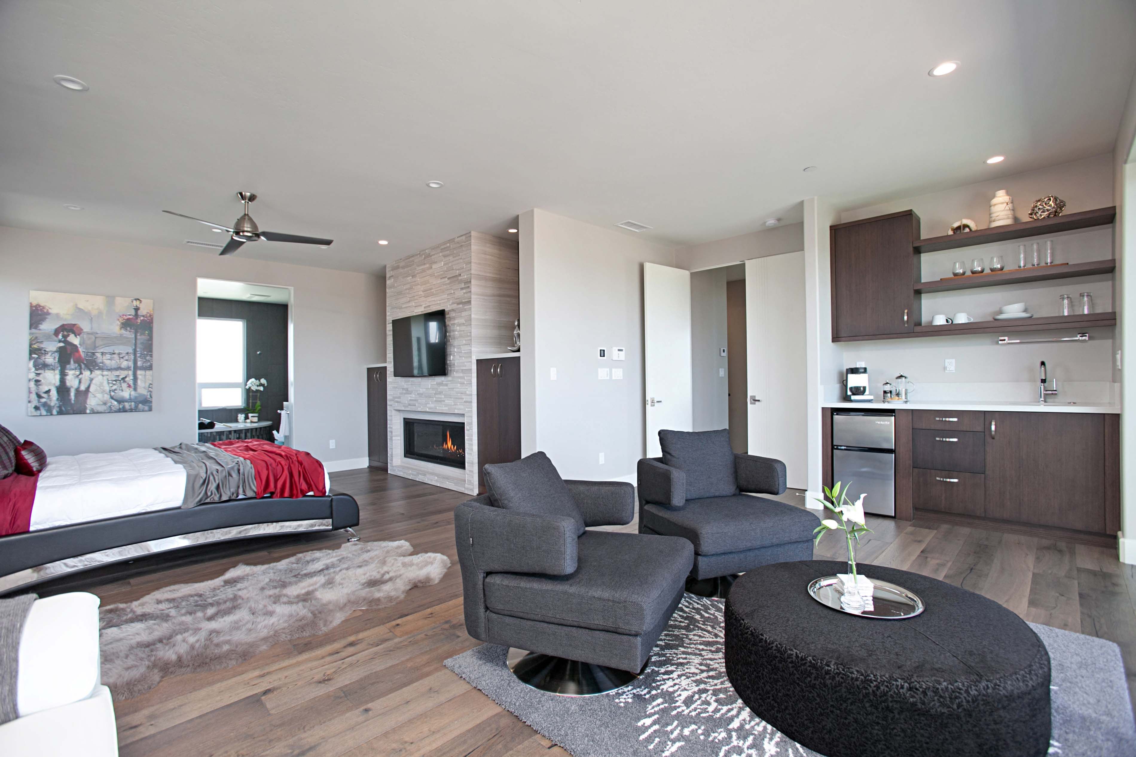 Open-concept living space with a bed on the left, two gray armchairs around a round black ottoman, and a shag rug. A stone fireplace with a mounted TV sits center while dark wood kitchenette with open shelves is to the right.