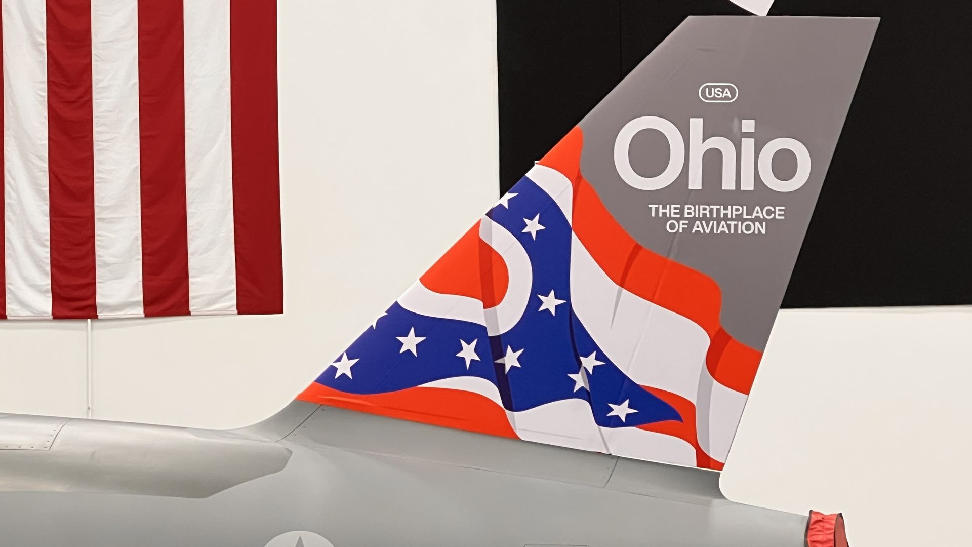 Exhibit scene: left shows a tall American flag; right features a gray aircraft tail with USA and Ohio and the title "The Birthplace of Aviation" on a black wall; red, white, and blue fabric drapes over the tail.