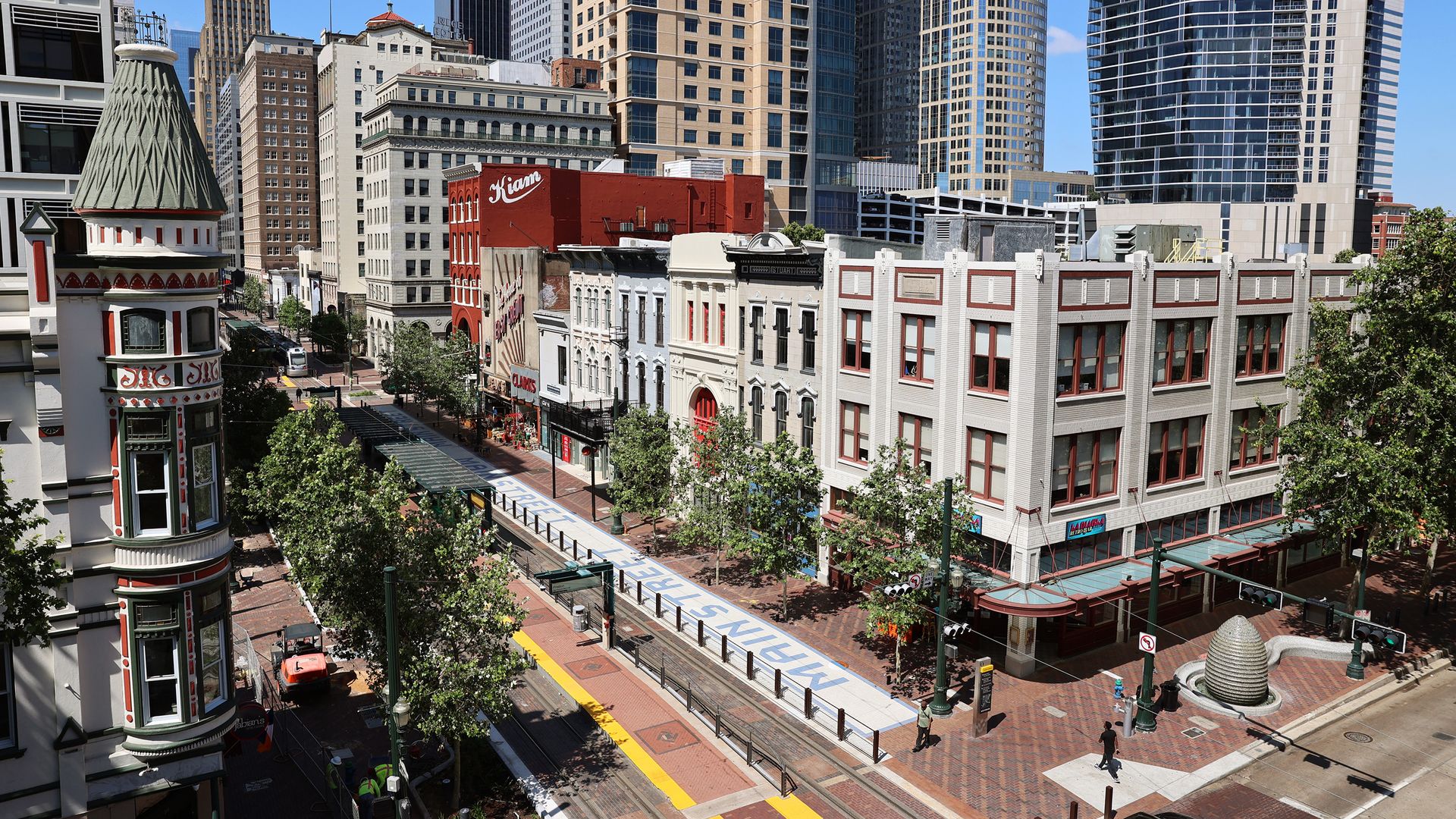 City skyline showing historic low-rise brick buildings in front and modern glass towers behind. A cone-topped green-tiled turret on the left; brick street with trees runs through the foreground.