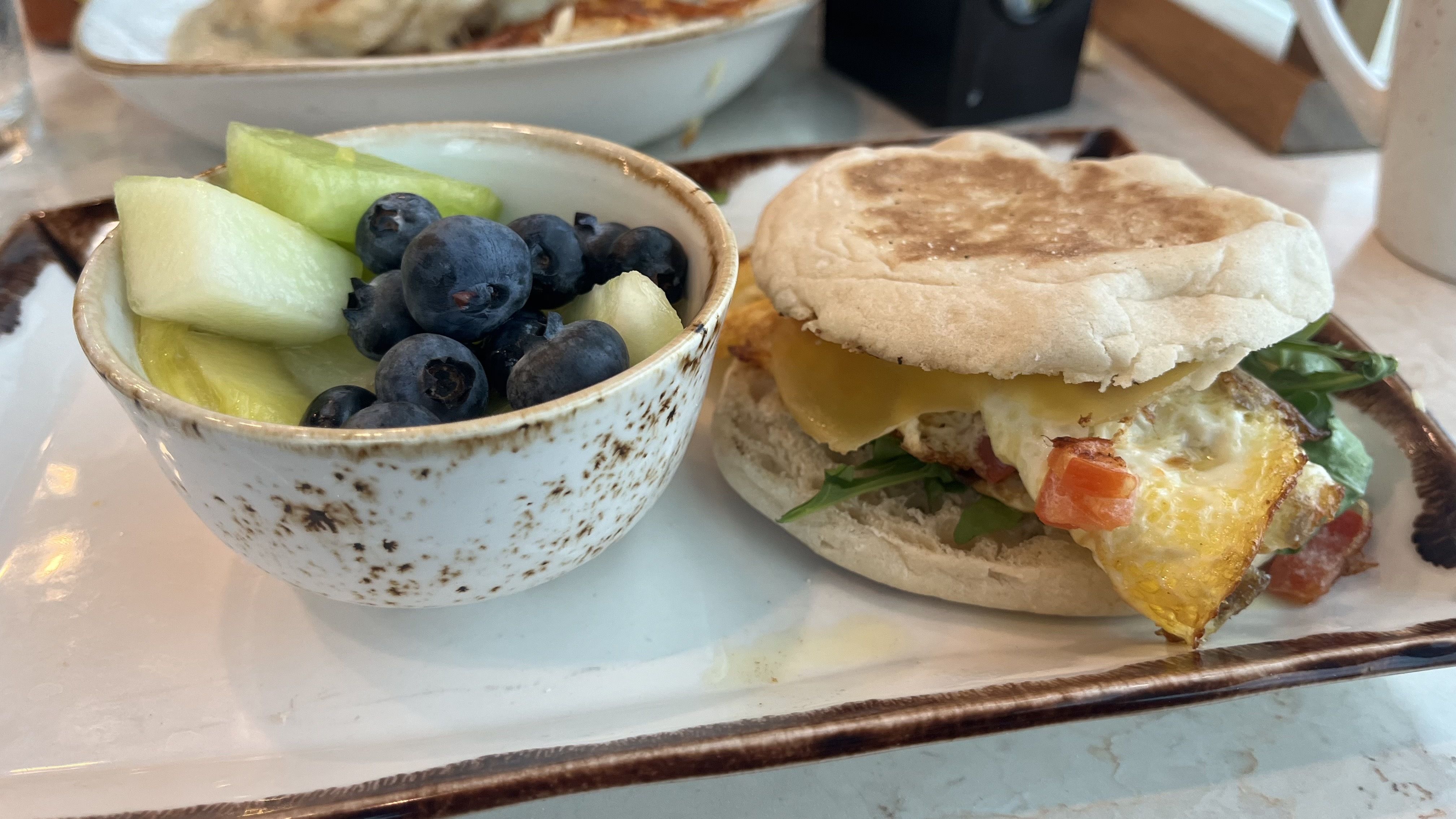 Plate with a small bowl of honeydew melon chunks and blueberries, next to a breakfast sandwich with fried egg, cheddar cheese, and greens on an English muffin.
