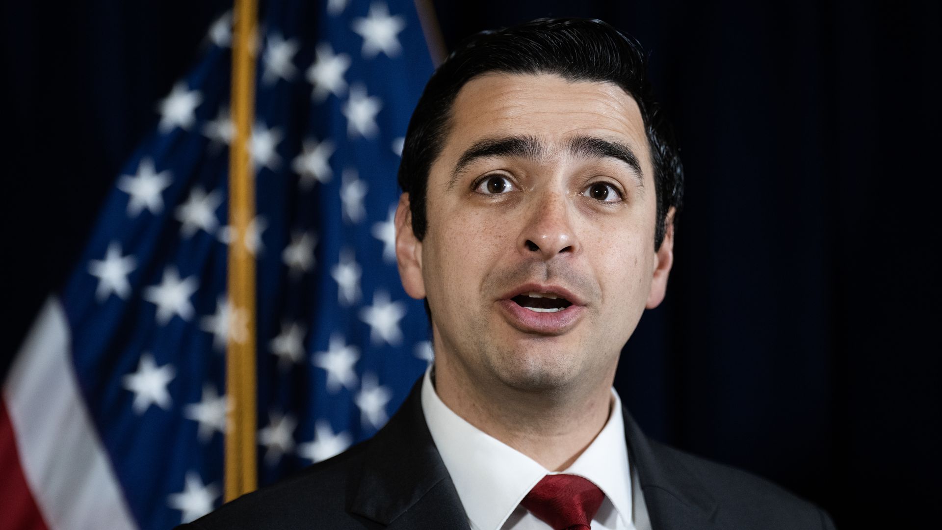 A man in a suit and necktie looks on while speaking. 