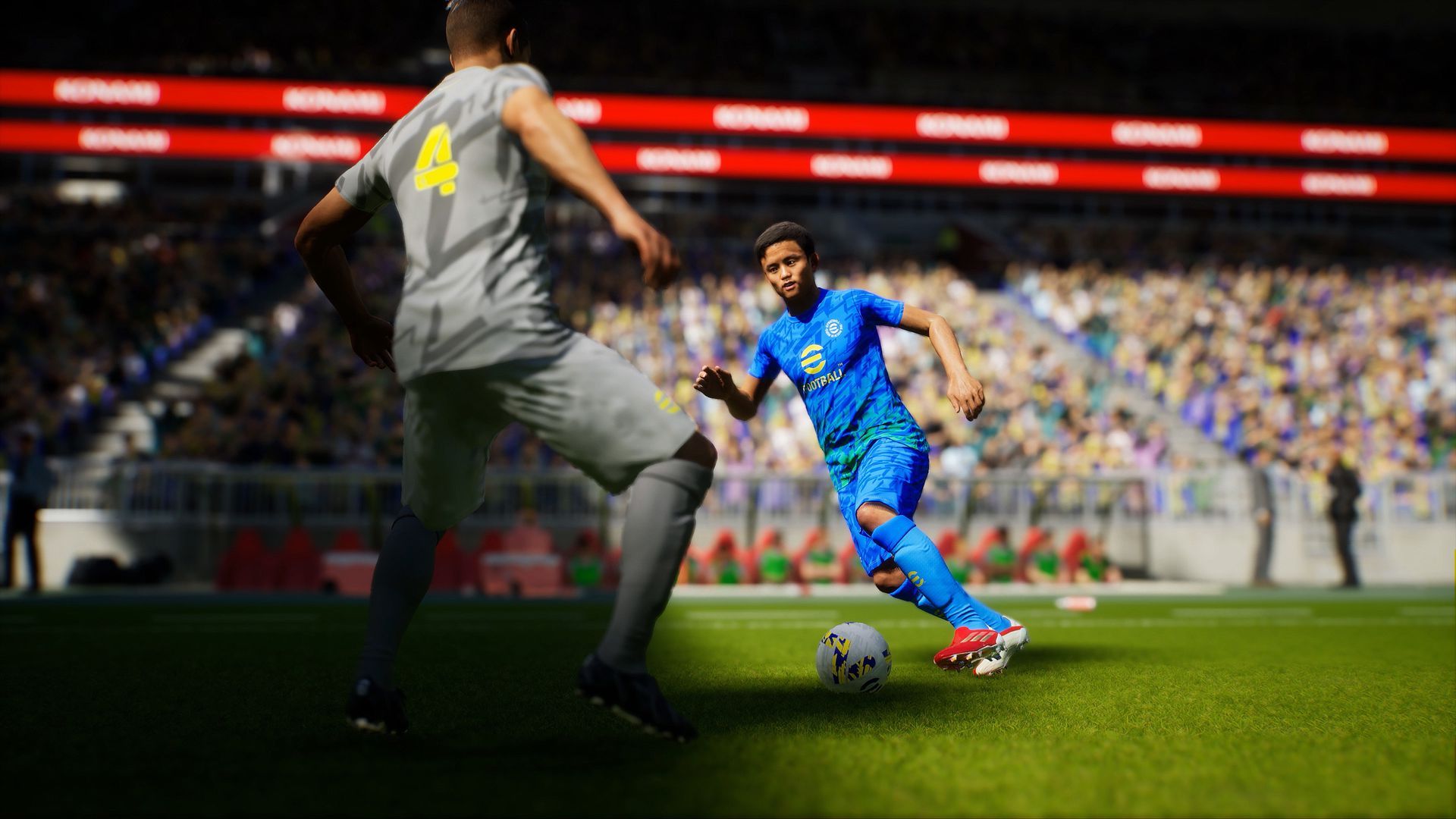 Screenshot of a scene from the video game eFootball 22, which shows one soccer playing in a blue uniform dribbling the ball as an opponent in grey approaches
