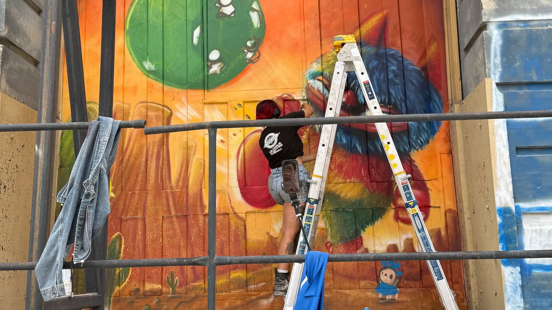 A mural painter on a metal ladder works on a vivid orange wall, adding color to a large graffiti piece; a denim jacket hangs on a railing nearby.