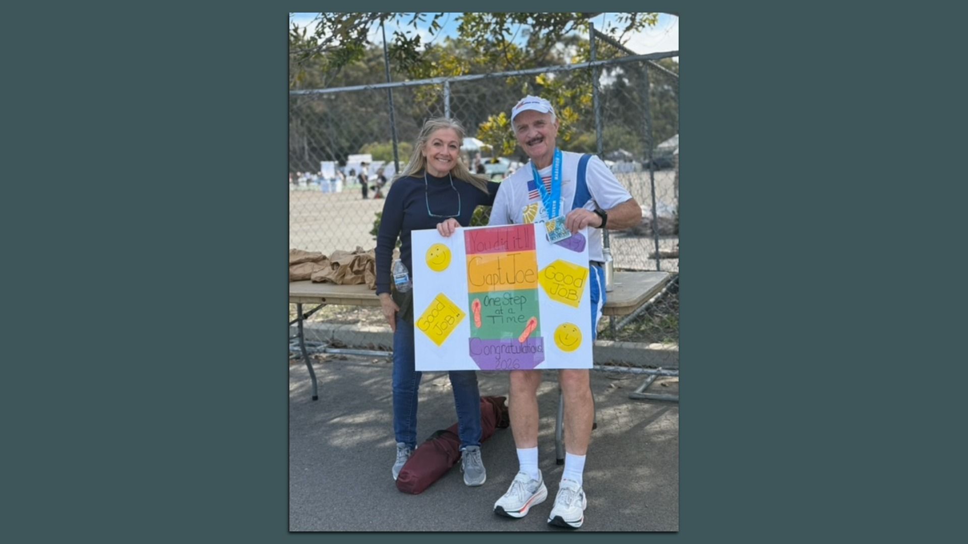 Smiling man with a medal and a woman hold a colorful congratulatory poster outdoors by a chain-link fence. The sign reads, "You did it!! Capt Joe, One step at a time, Congratulations 2026."