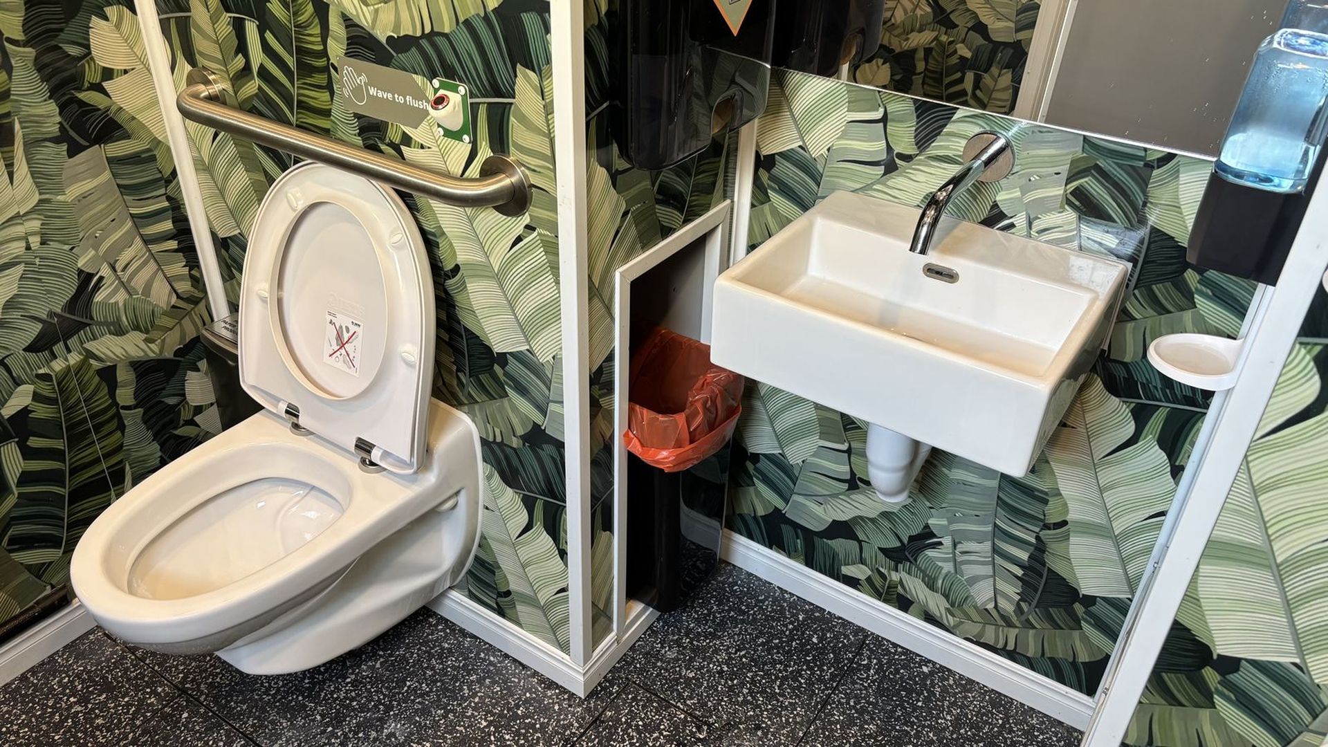 A photo of a toilet with the seat up and a wash basin next to it. The walls are covered in palm leaf wallpaper.