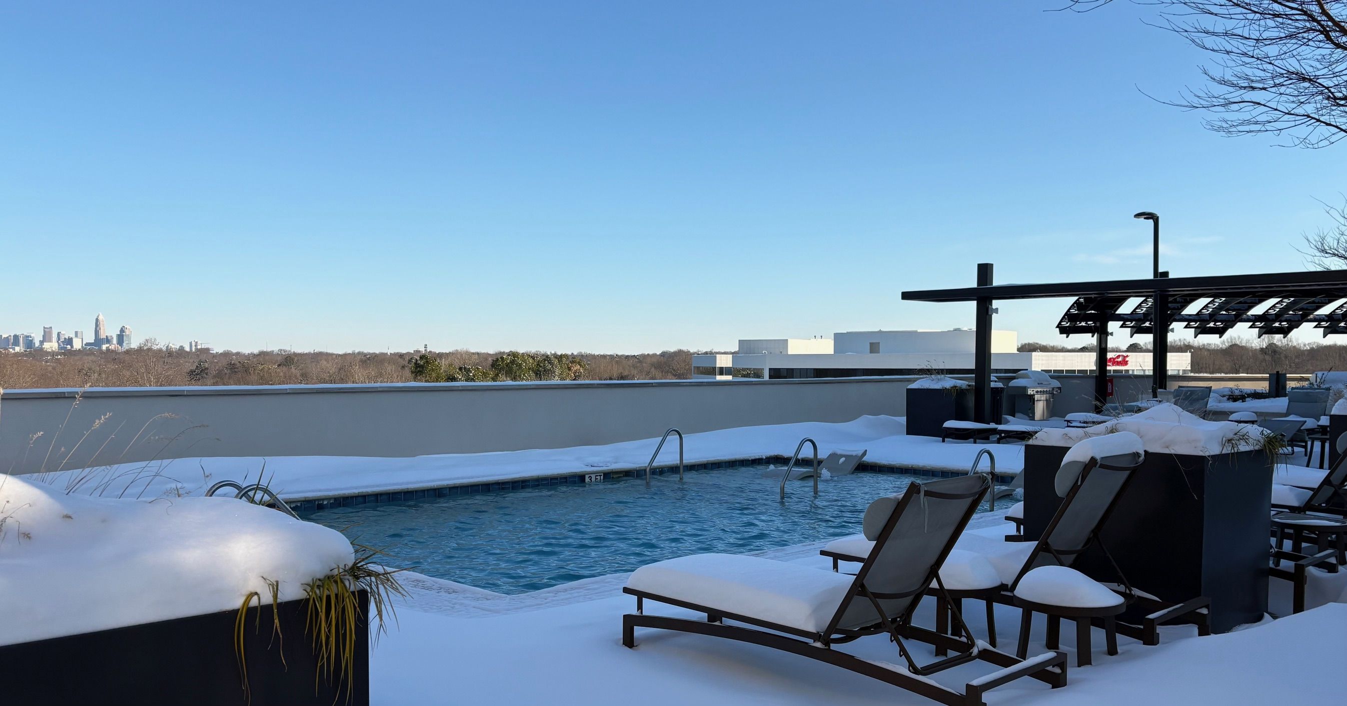 Outdoor rooftop pool surrounded by snow-covered lounge chairs and plants, with a clear blue sky and a distant city skyline visible over a wall in the background.