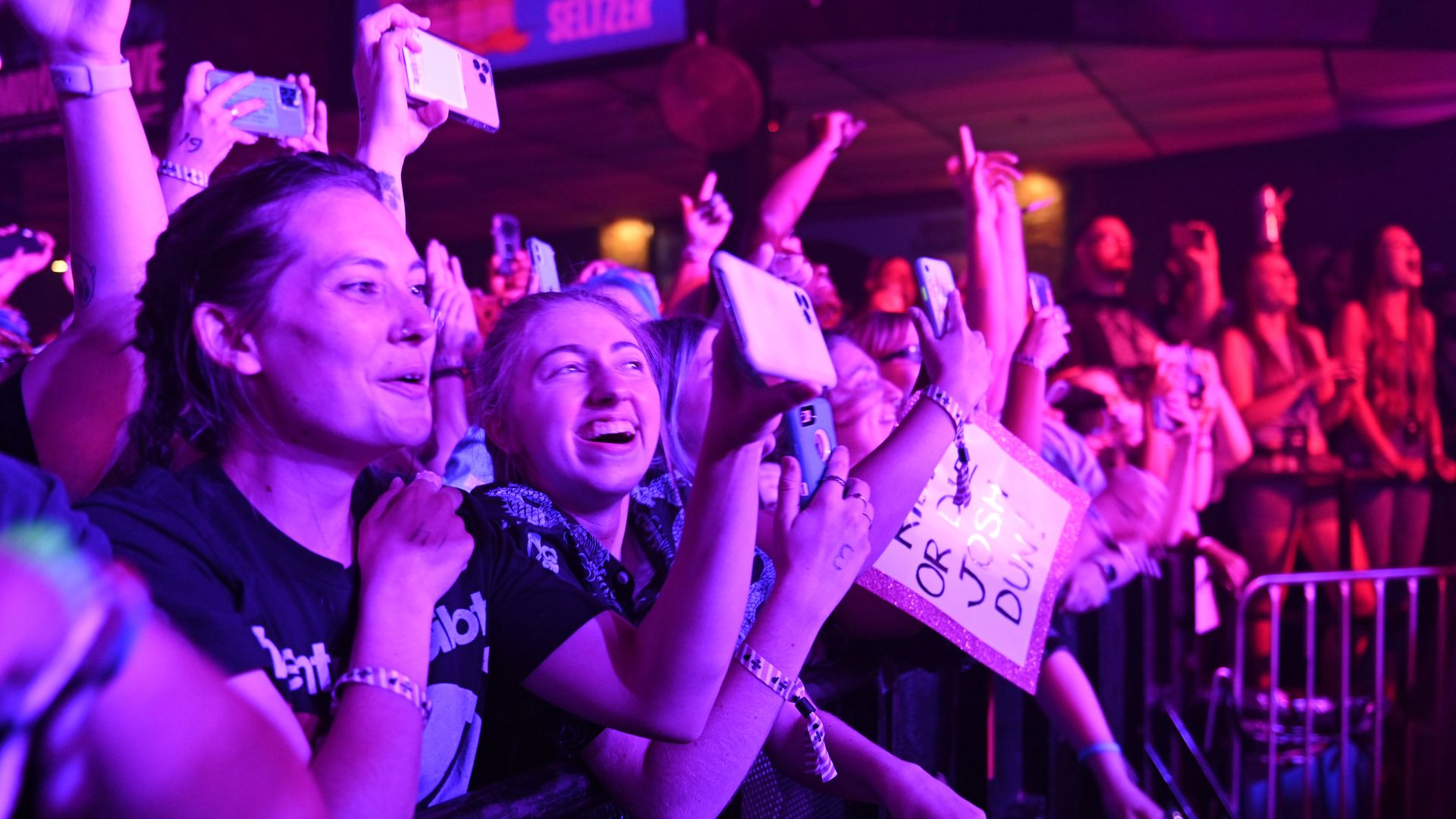 A crowd hangs on the barrier at a concert while holding cell phones and signs