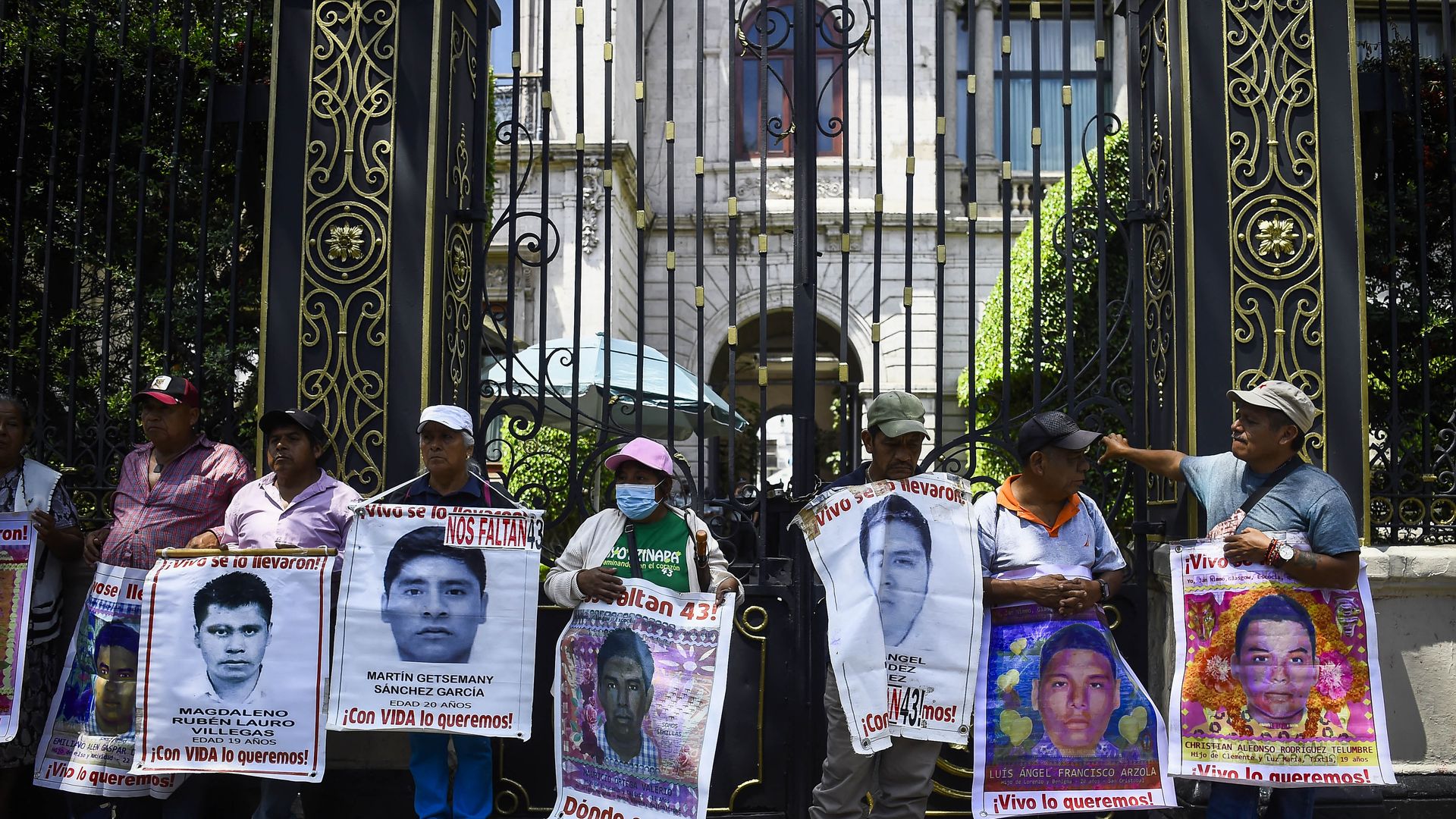 eight people are shown holding large signs with photos of young disappeared men. the people are standing in front Mexico's Senate building which has large black and golden gates. 