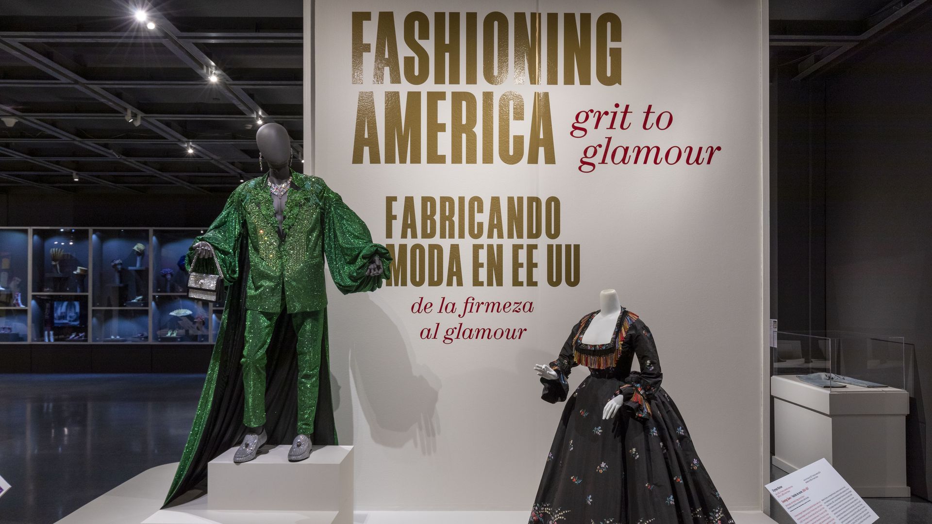 Two mannequins are dressed in front of a panel that reads "Fashioning America: Grit to Glamour." The mannequin at left wears a glitter green suit with a matching cape, and the one at right wears an 1860s style gown with a low neckline and bell skirt.