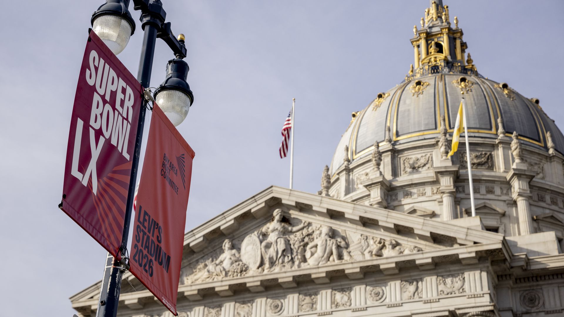 A red Super Bowl LX banner and a Levi's Stadium 2026 sign hang on a lamp post near a grand historic building with a dome topped by gold details and flags against a clear sky.
