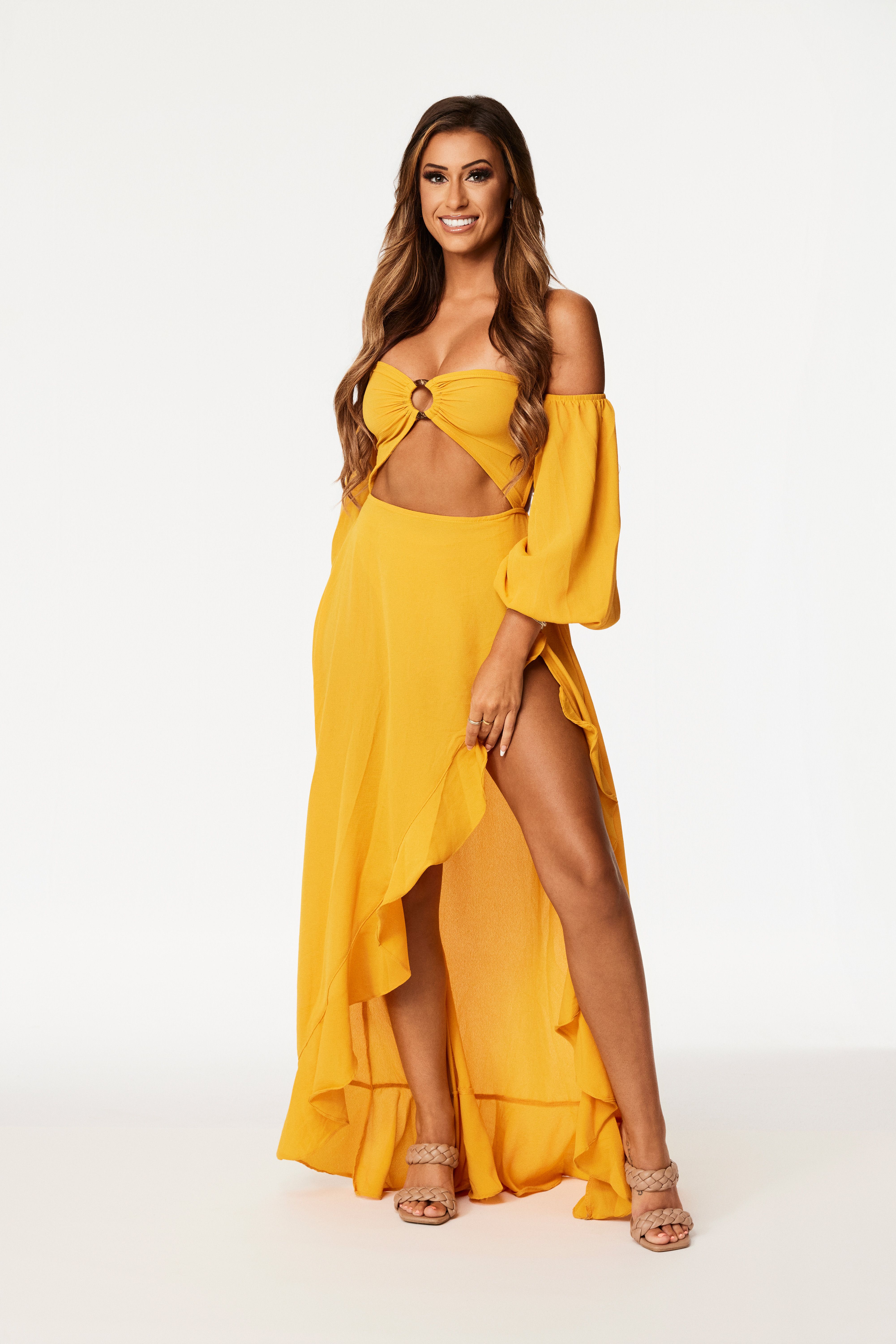 Woman with long brown hair, wearing a yellow off-the-shoulder top with ring detail and matching long skirt with high slit, beige braided strap heels, smiling against white background.