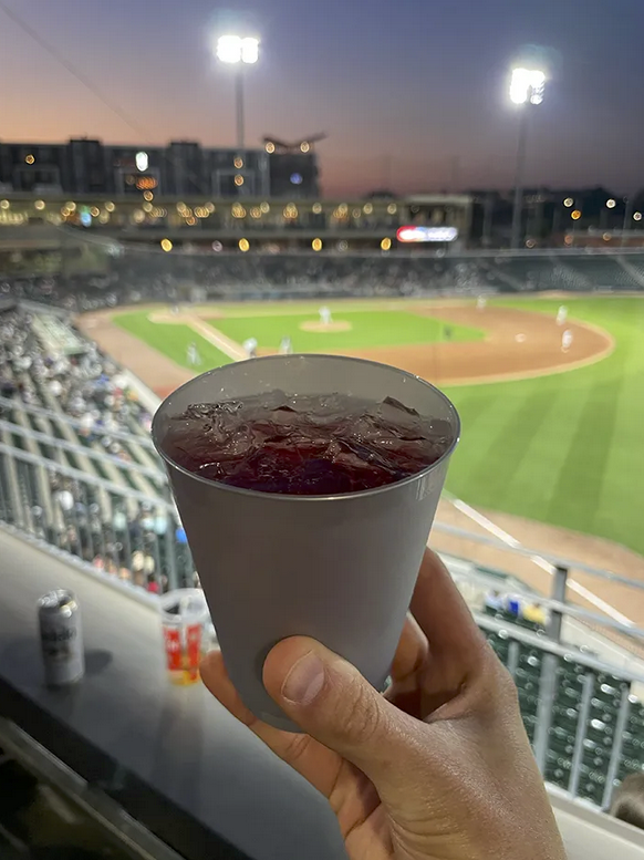 A plastic cup holding a red drink in front of a baseball stadium field. 