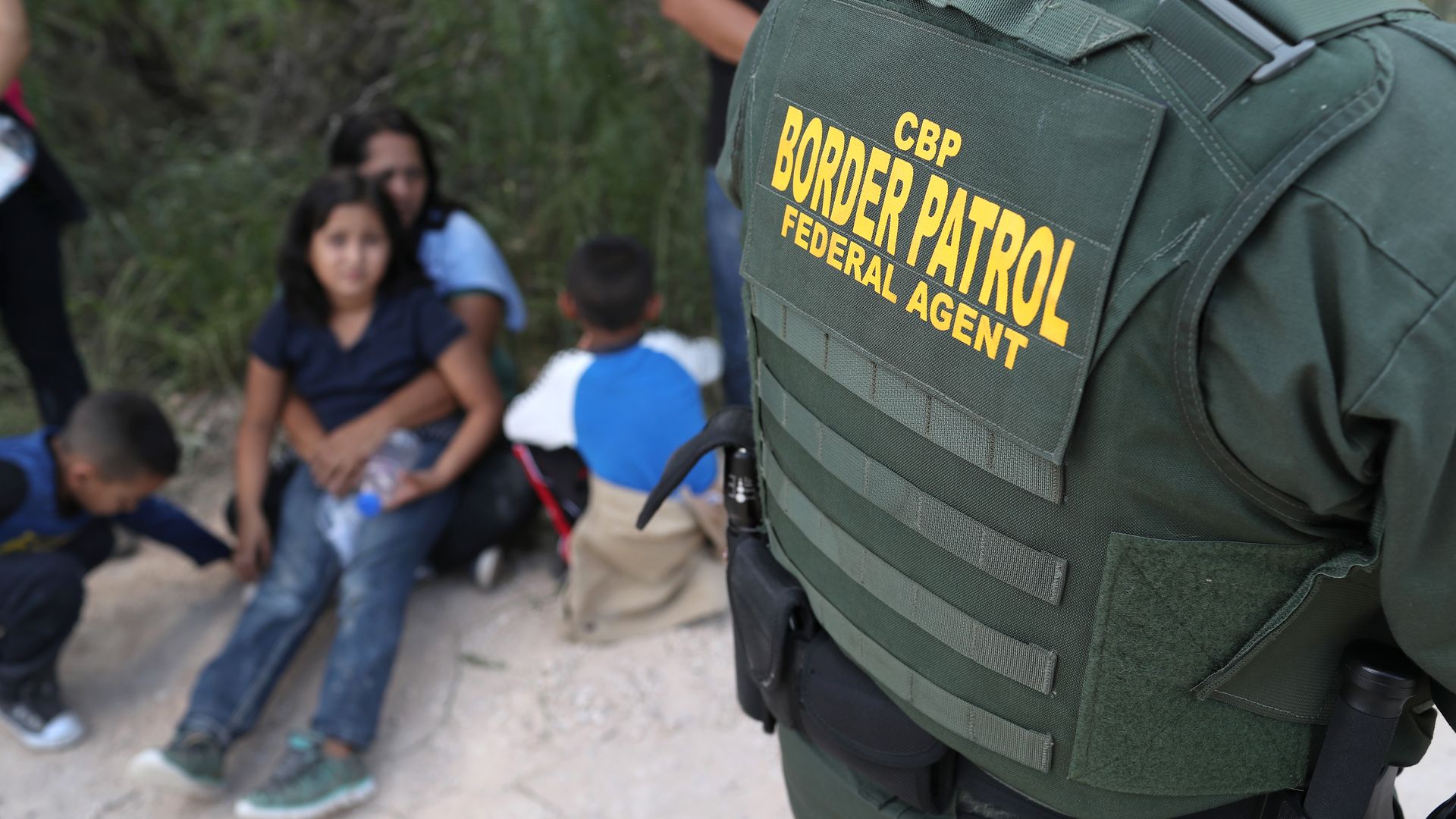 Border Patrol agent jacket with arrested immigrants sitting in the sand in the background