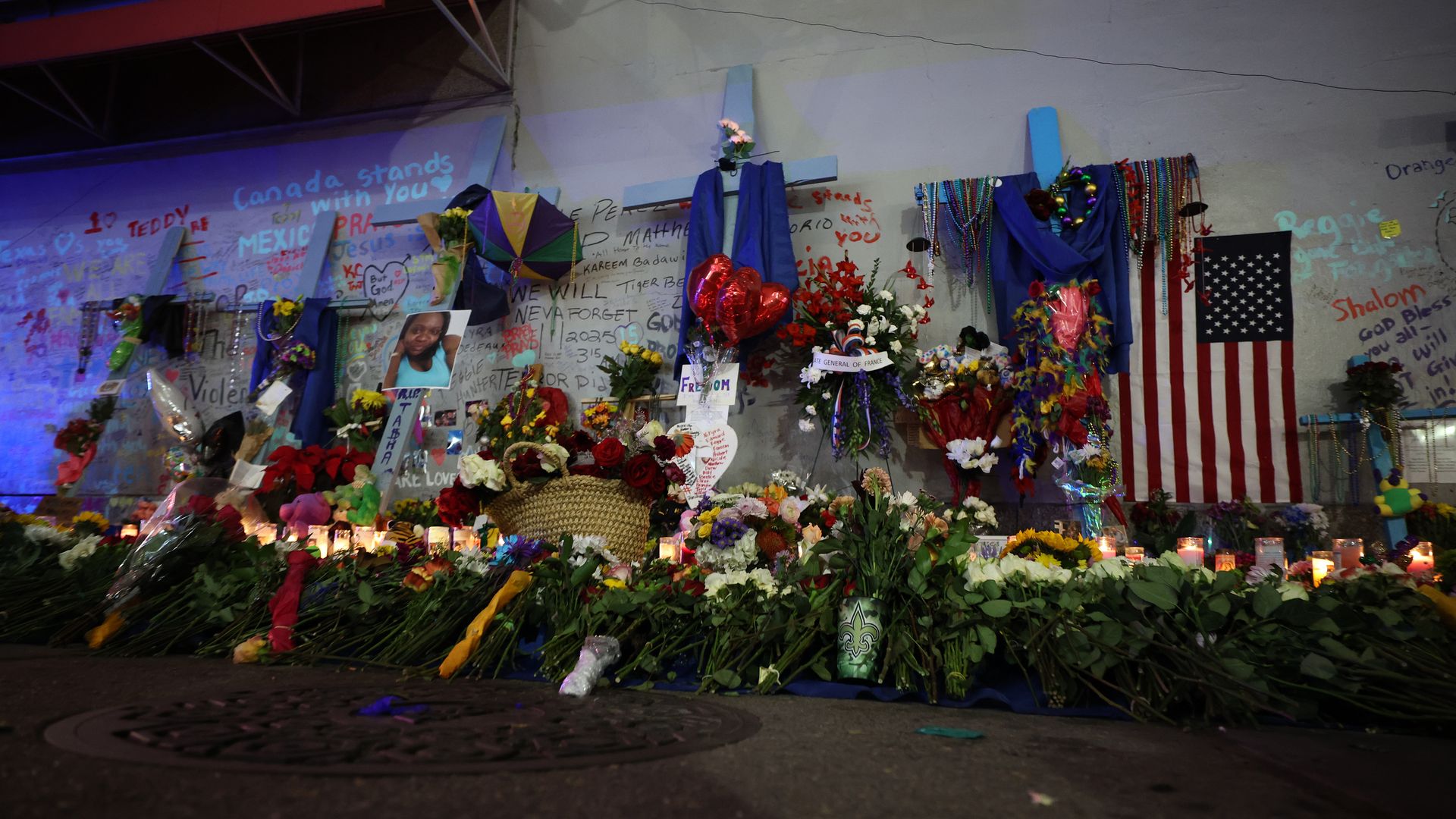 A gray wall is covered with commemorative graffiti, bouquets, crosses and other memorial tokens. 