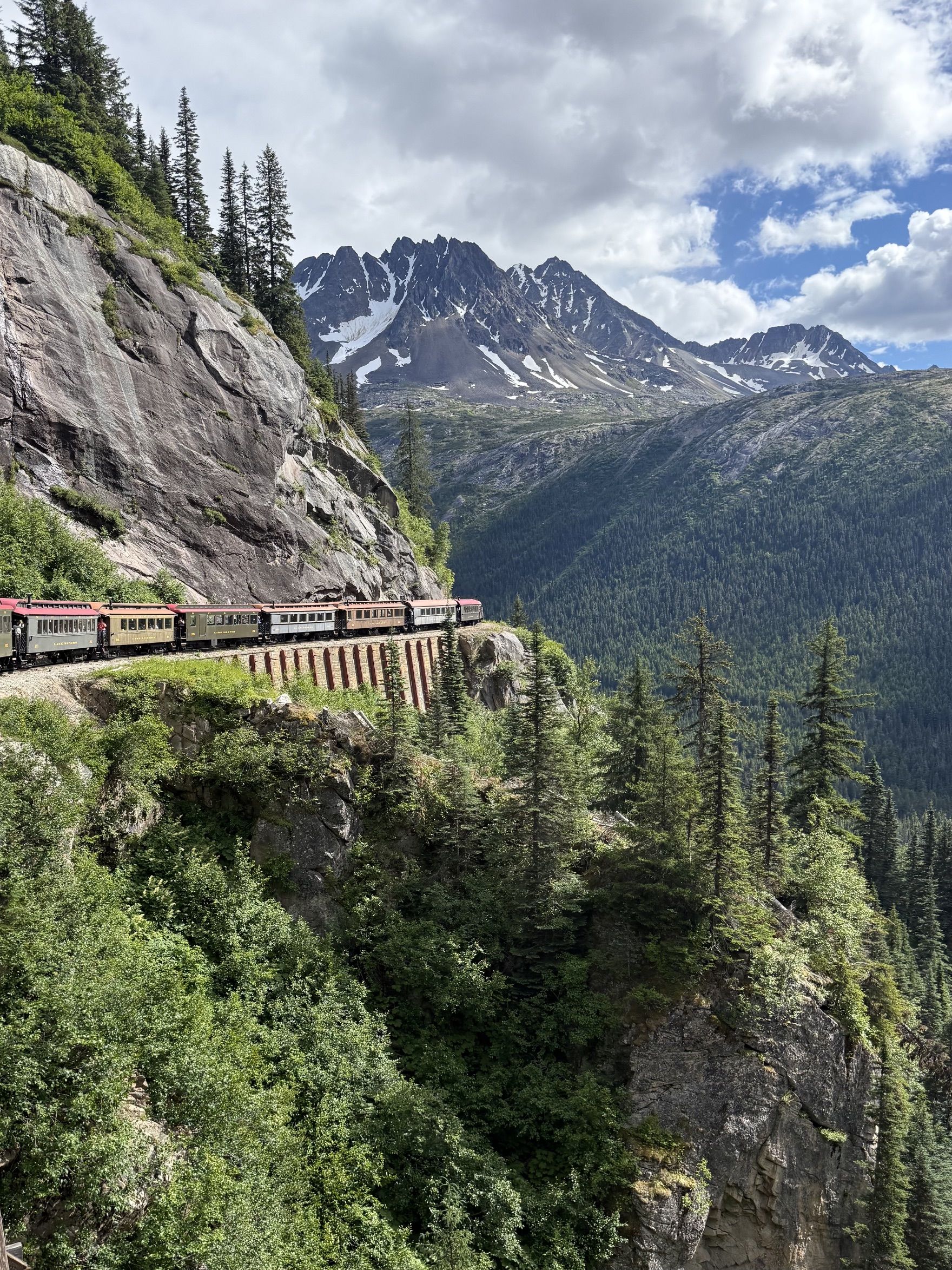 A colorful passenger train winds along a rocky cliff, with evergreen trees, snow-dusted mountains in the distance, and a bright, partly cloudy sky.