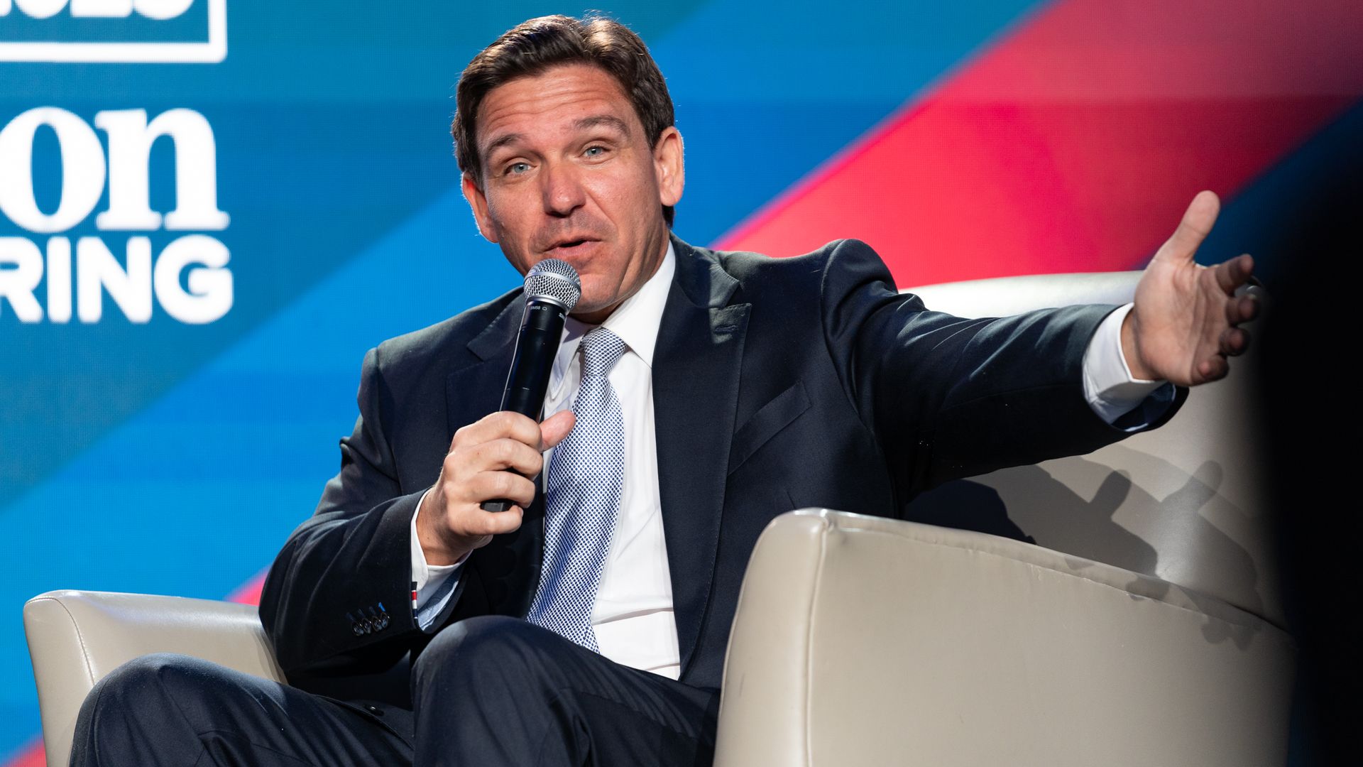 Ron DeSantis, in a navy blue suit and light blue tie, sits in a chair, speaking into a microphone.