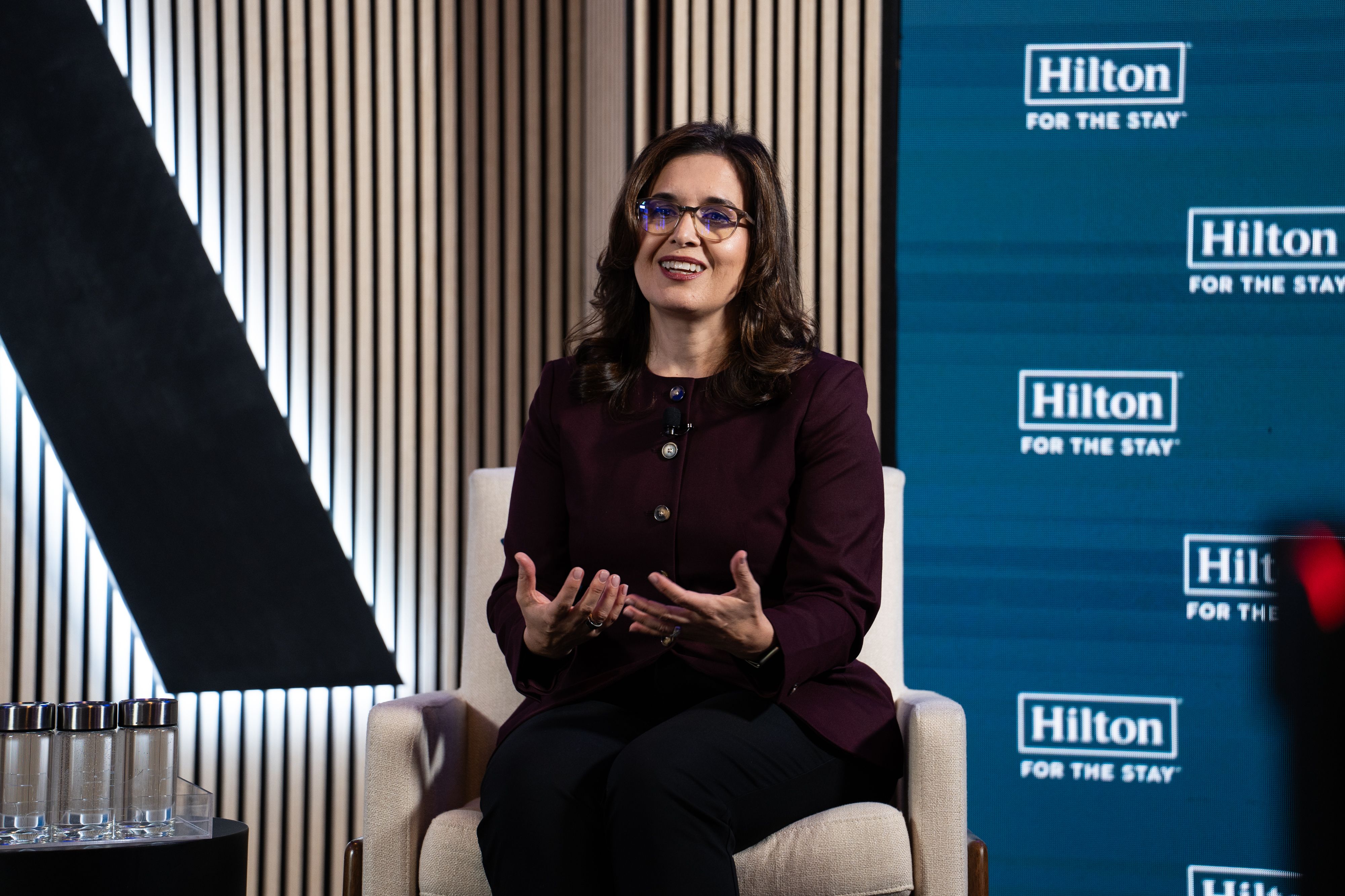 Smiling woman with glasses in a burgundy top sits in a beige chair, gesturing as she speaks. Behind her is a blue backdrop displaying Hilton logos reading "Hilton FOR THE STAY".