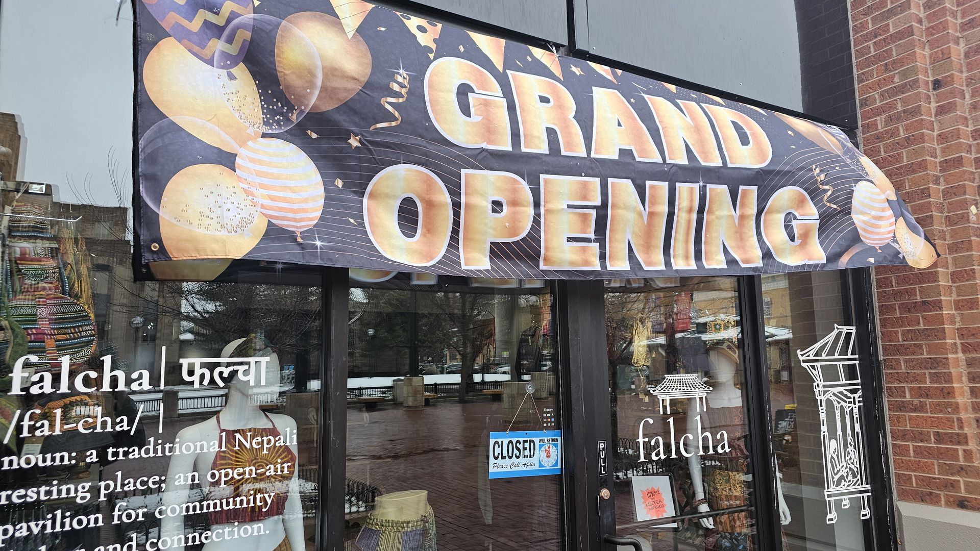 Storefront with a dark banner reading "GRAND OPENING" in bold orange-gold letters, decorated with balloons and confetti; brick wall on the right, glass doors, and street reflections.
