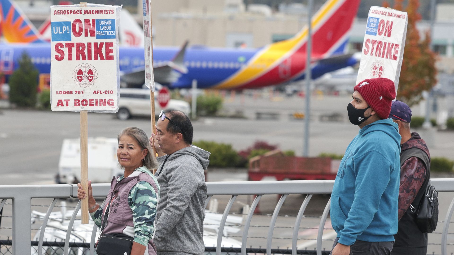 Two people hold signs that say "on strike" in front of airplanes in the background, with two other people joining them at the picket line.