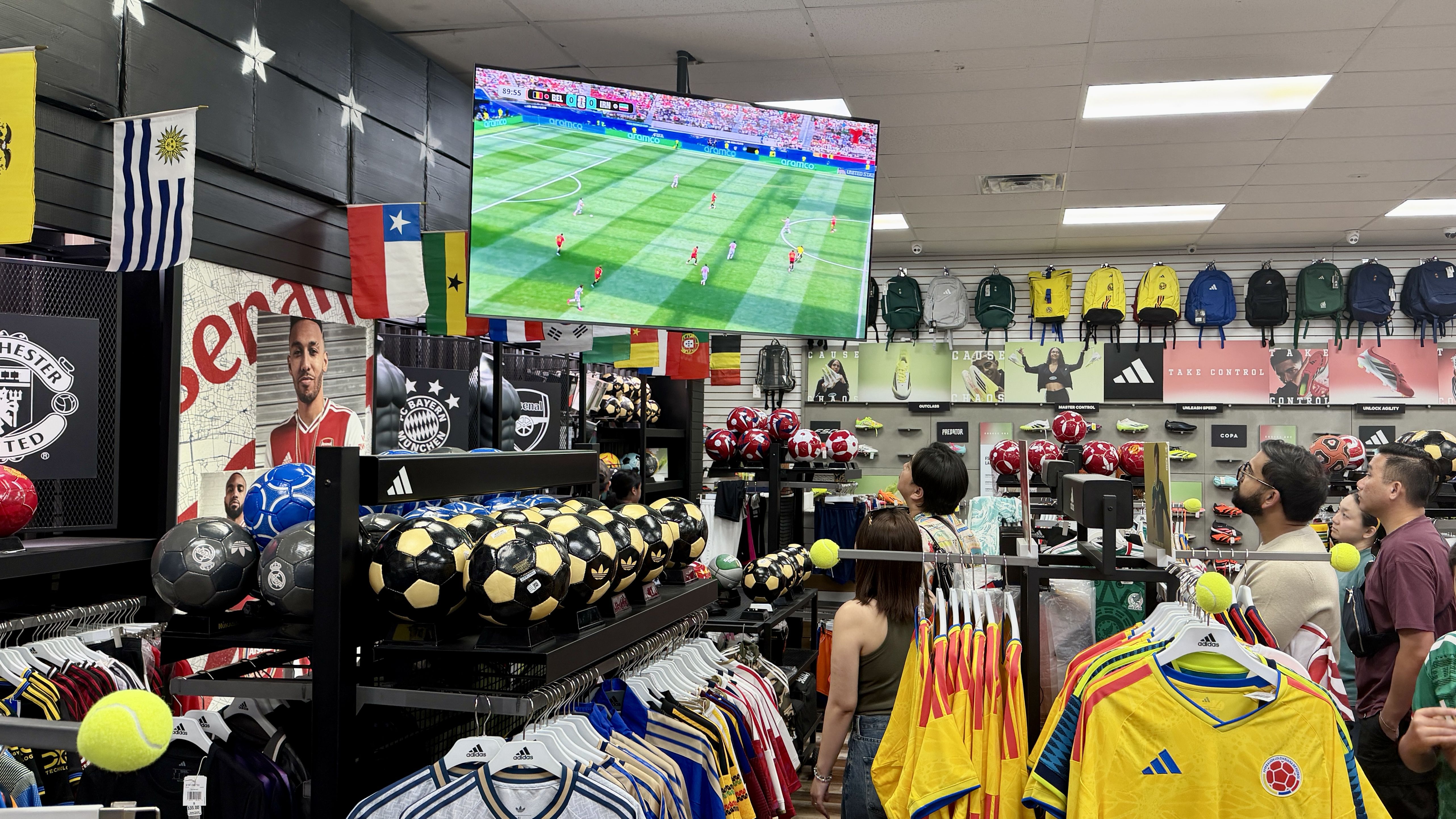 Inside a sports store, a large screen shows a soccer match while shoppers watch. Flags hang from the ceiling; shelves brim with colorful jerseys, backpacks, and soccer balls.