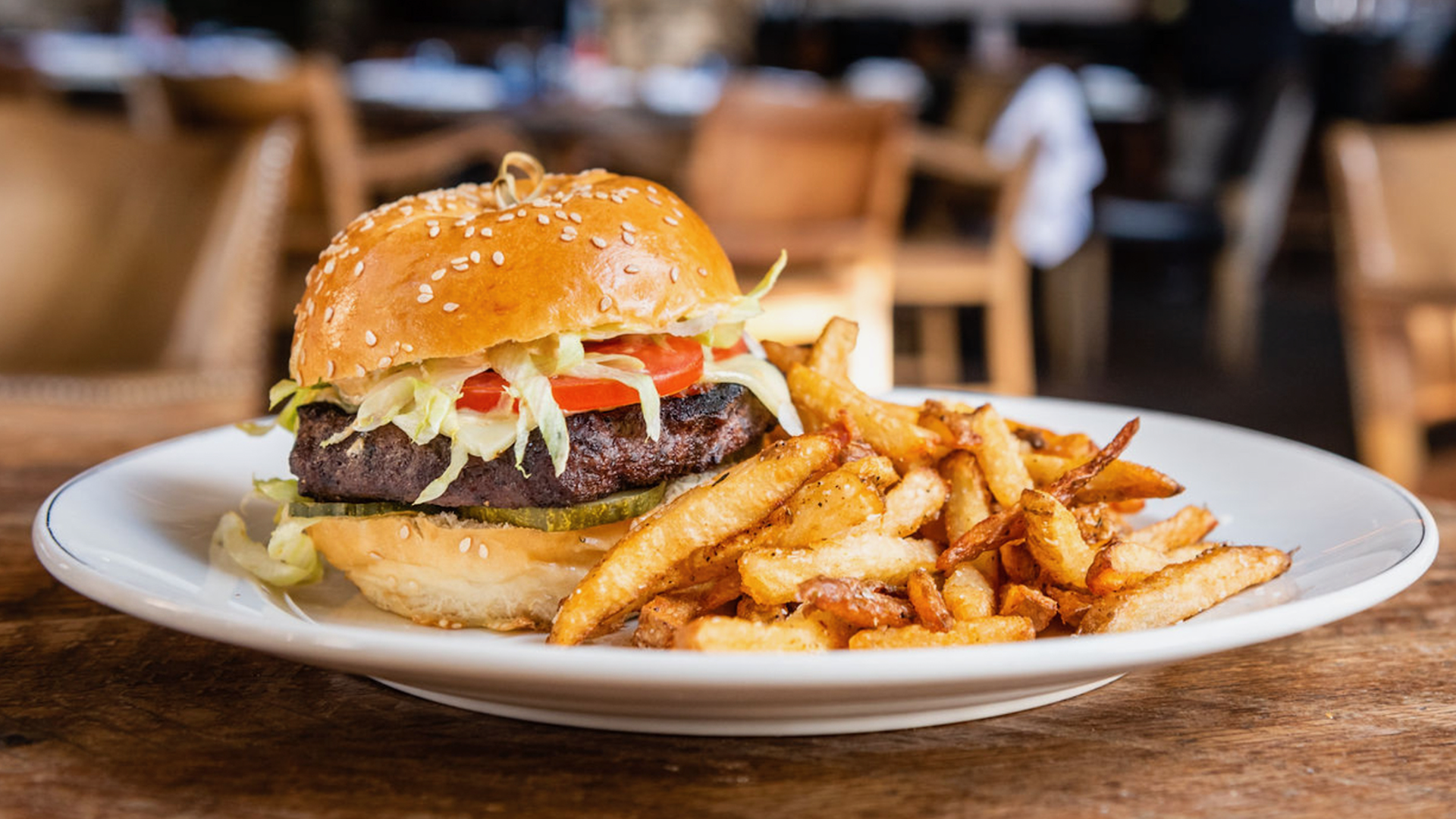 Sesame-seed bun hamburger with lettuce, tomato, pickles and a beef patty on a white plate, with a generous side of golden fries in a cozy restaurant.