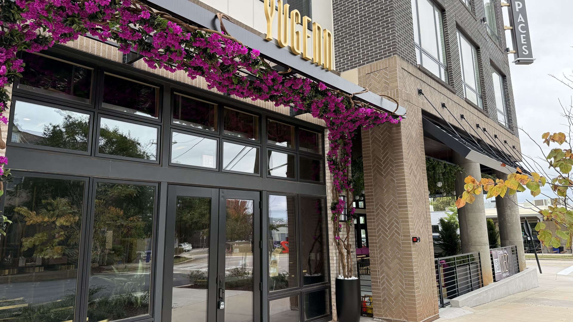 Entrance of a modern building with a sign reading YUGENI, decorated with purple flowers above black-framed glass doors. Nearby building has a sign that says PACES.