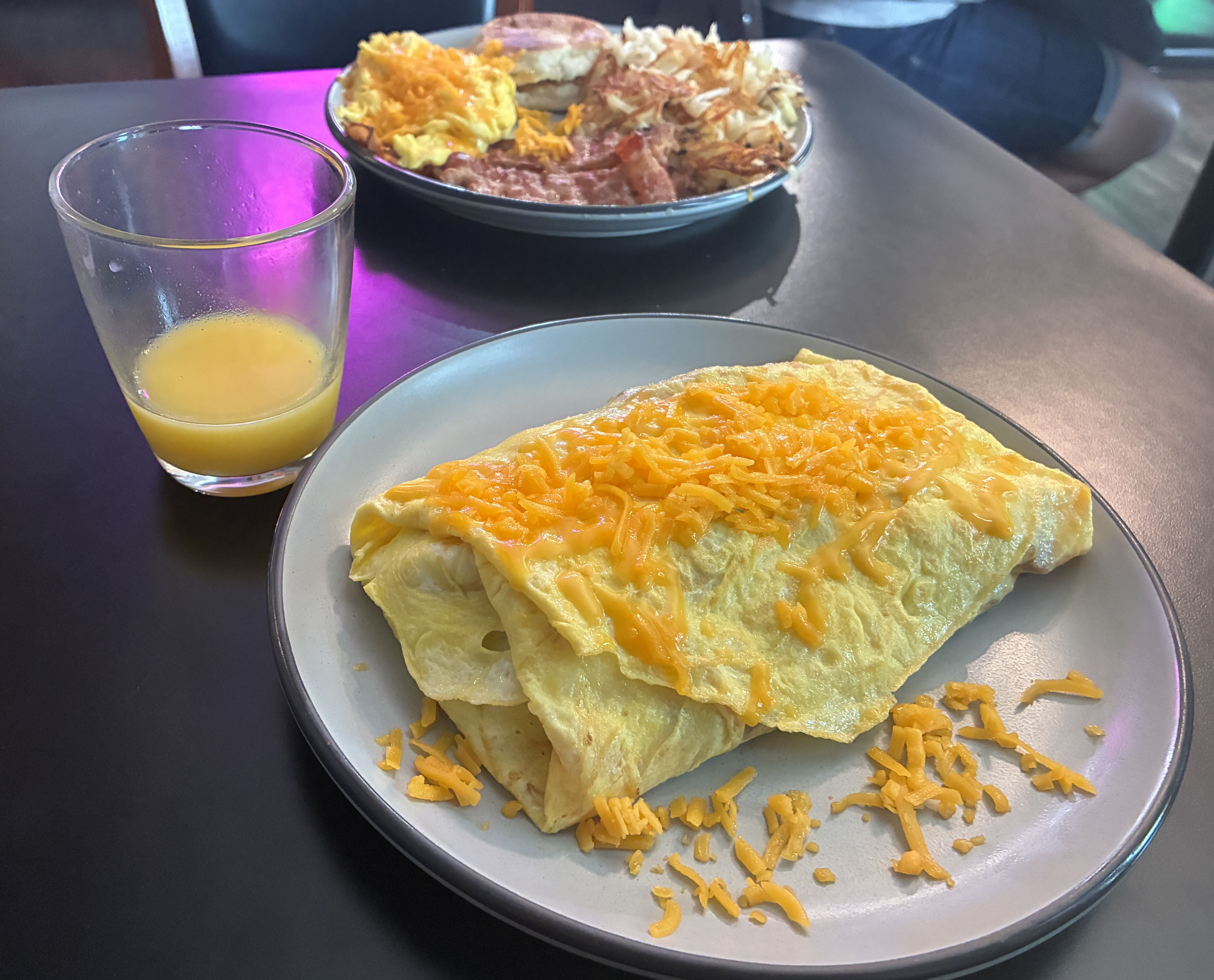Two plates on a dark table: foreground shows a cheesy folded flatbread topped with shredded cheddar; background features a hash-like dish. A glass of orange juice sits to the left; purple glow reflects.