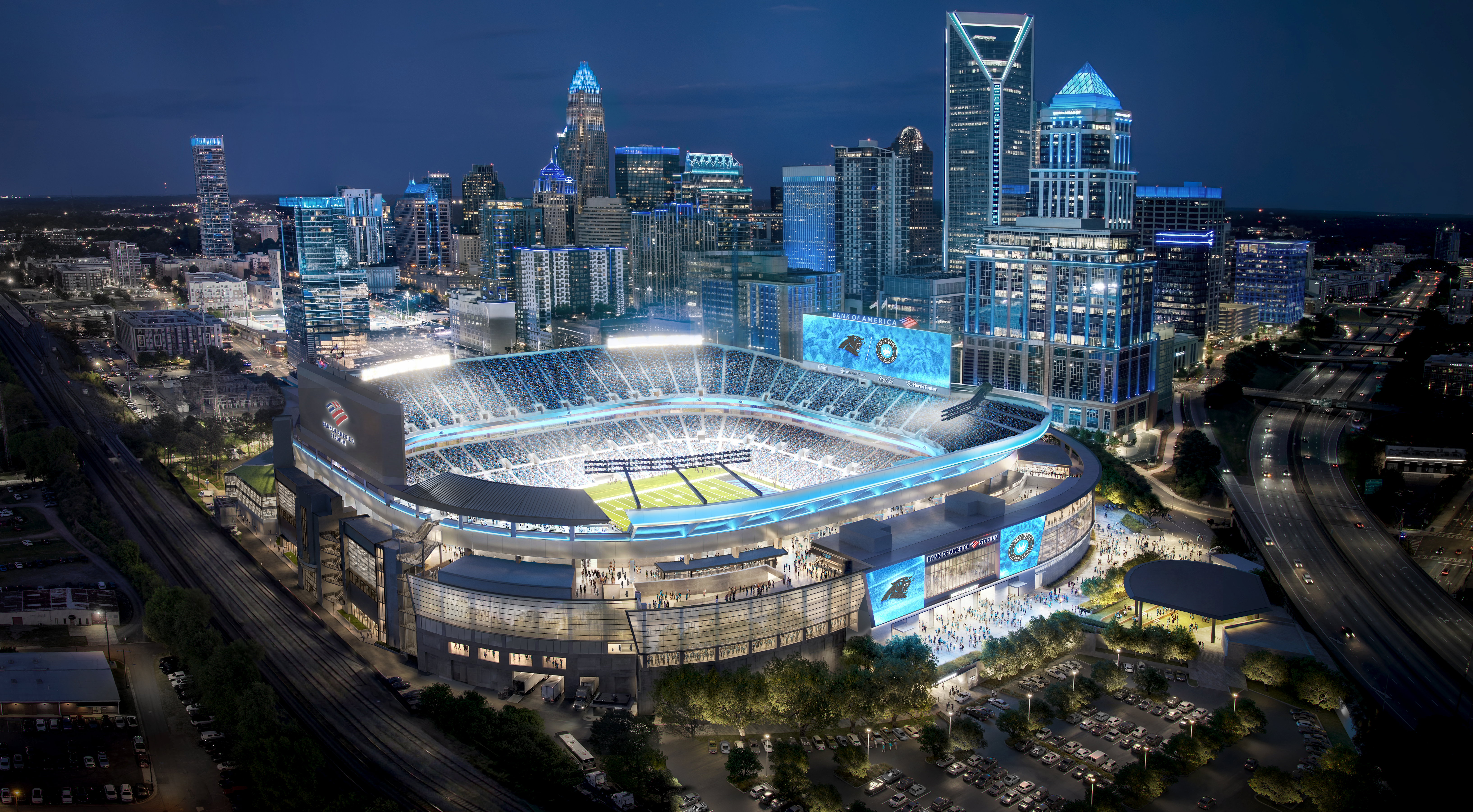 Night aerial view of a blue-lit city skyline with tall glass skyscrapers; in the foreground a large stadium with curved seating, a bright green field, and a prominent digital scoreboard.