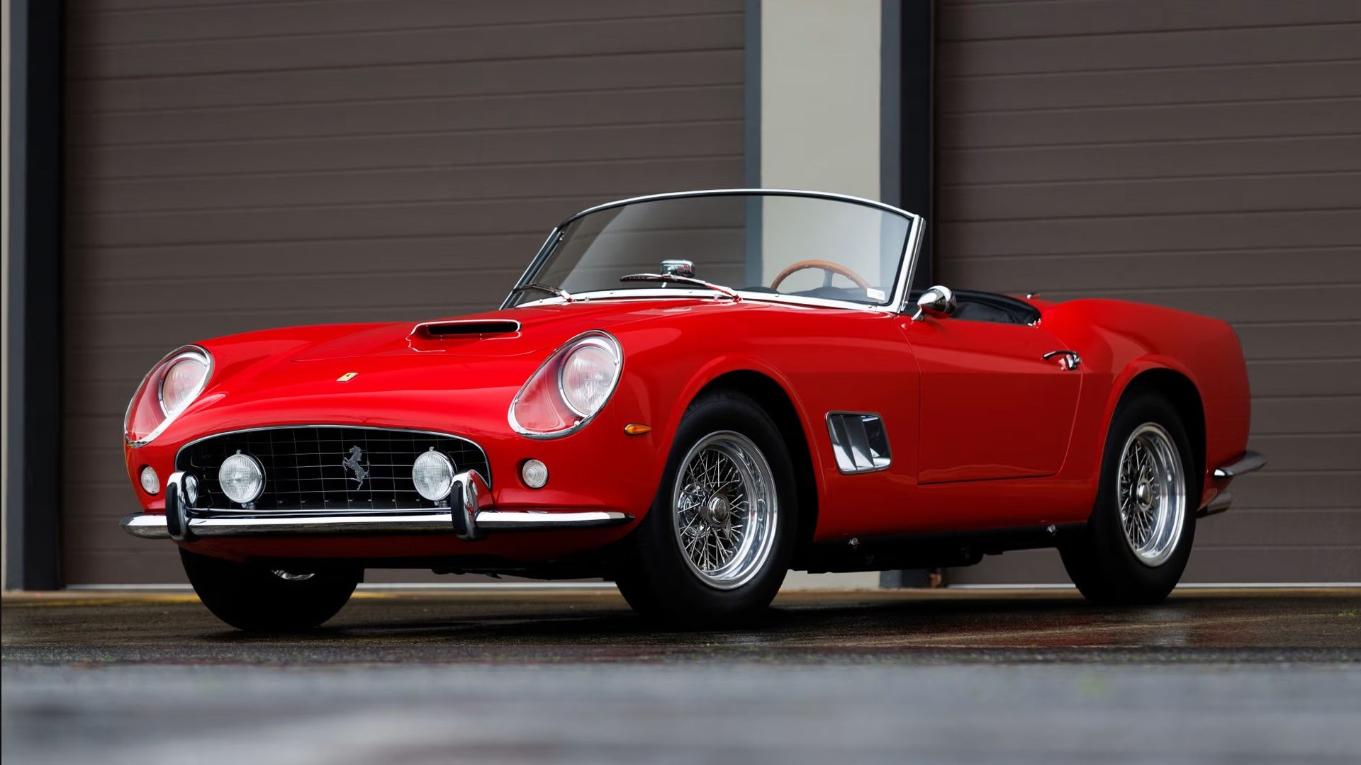 Red classic Ferrari convertible with top down, rounded headlights, chrome grille with the Ferrari emblem, chrome bumper, and wire-spoke wheels, parked in front of a brown garage door.