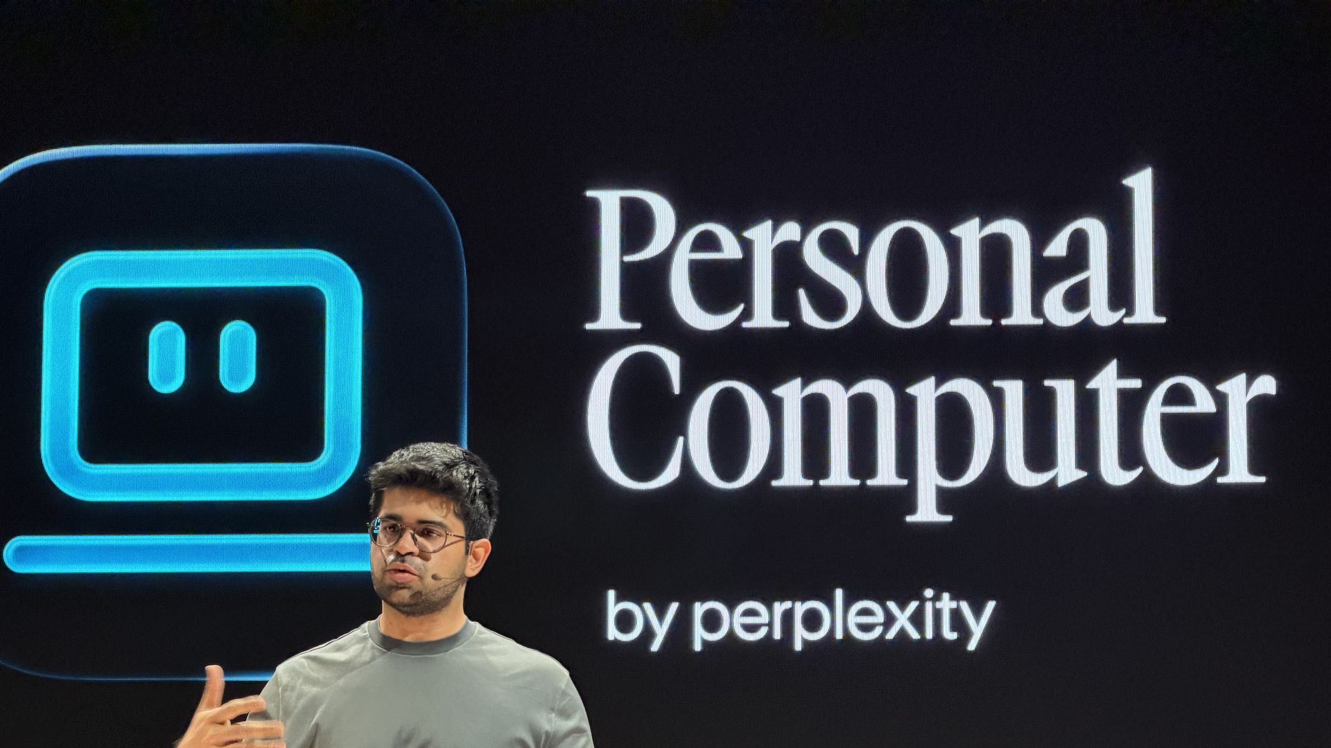 Speaker in a gray t-shirt presents on a stage with a large screen showing a blue rounded-square icon and the title "Personal Computer" by perplexity.