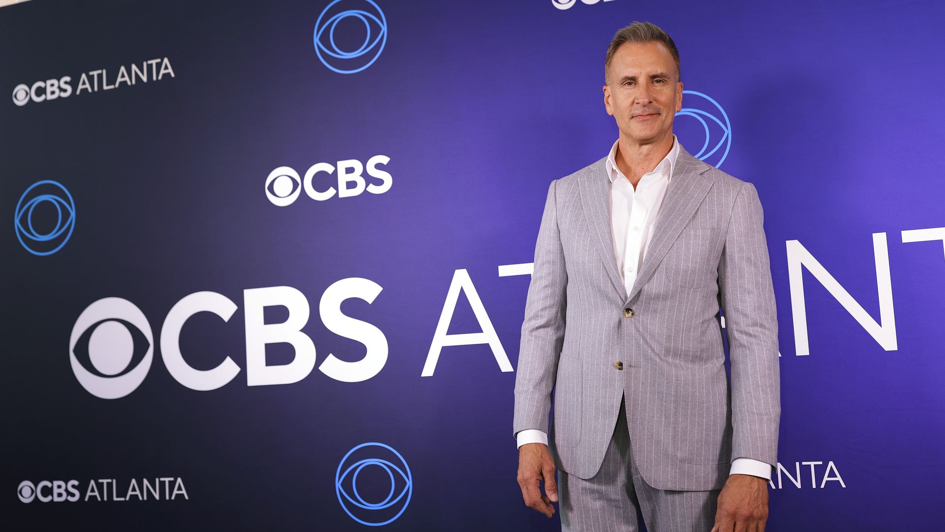 A man in a gray pin-striped suit and no tie stands in front of a purple screen that reads "CBS Atlanta" and has the CBS logo.