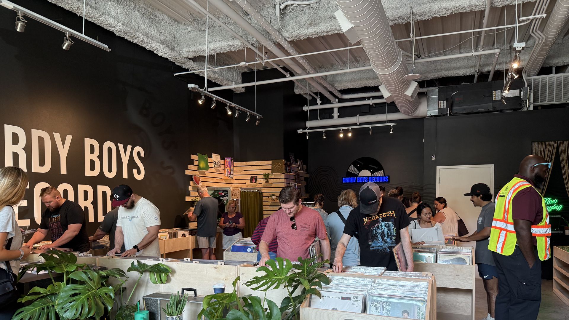 Interior of Hardy Boys Records store with people browsing vinyl records in wooden bins labeled New Arrivals. Black walls, plants, and industrial ceiling with exposed ducts.