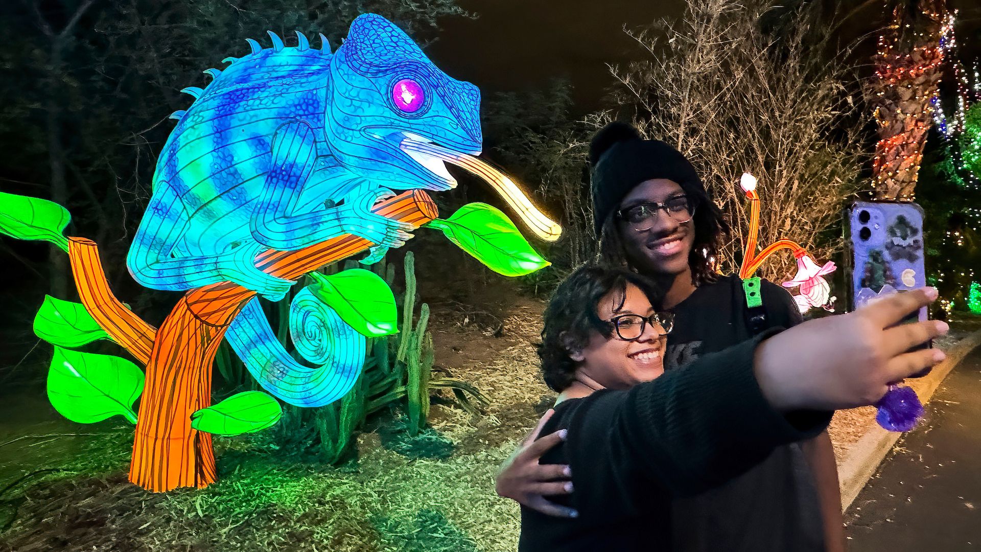 Two people smiling and taking a selfie at night next to a large, illuminated blue chameleon figure on an orange branch with green leaves, with bare trees and colorful lights in the background.