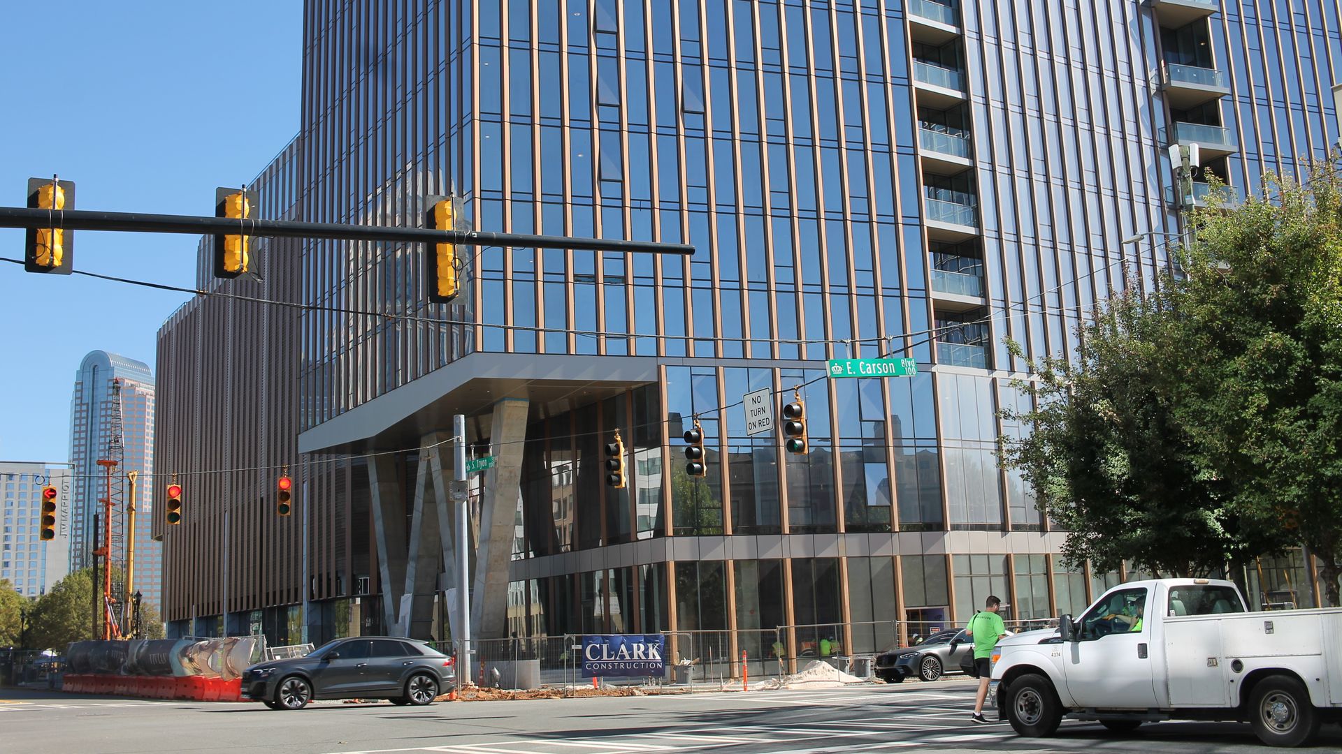 Modern glass building with vertical metallic stripes under blue sky at E. Carson Blvd intersection, featuring traffic lights, cars, construction sign, and a person crossing the street.
