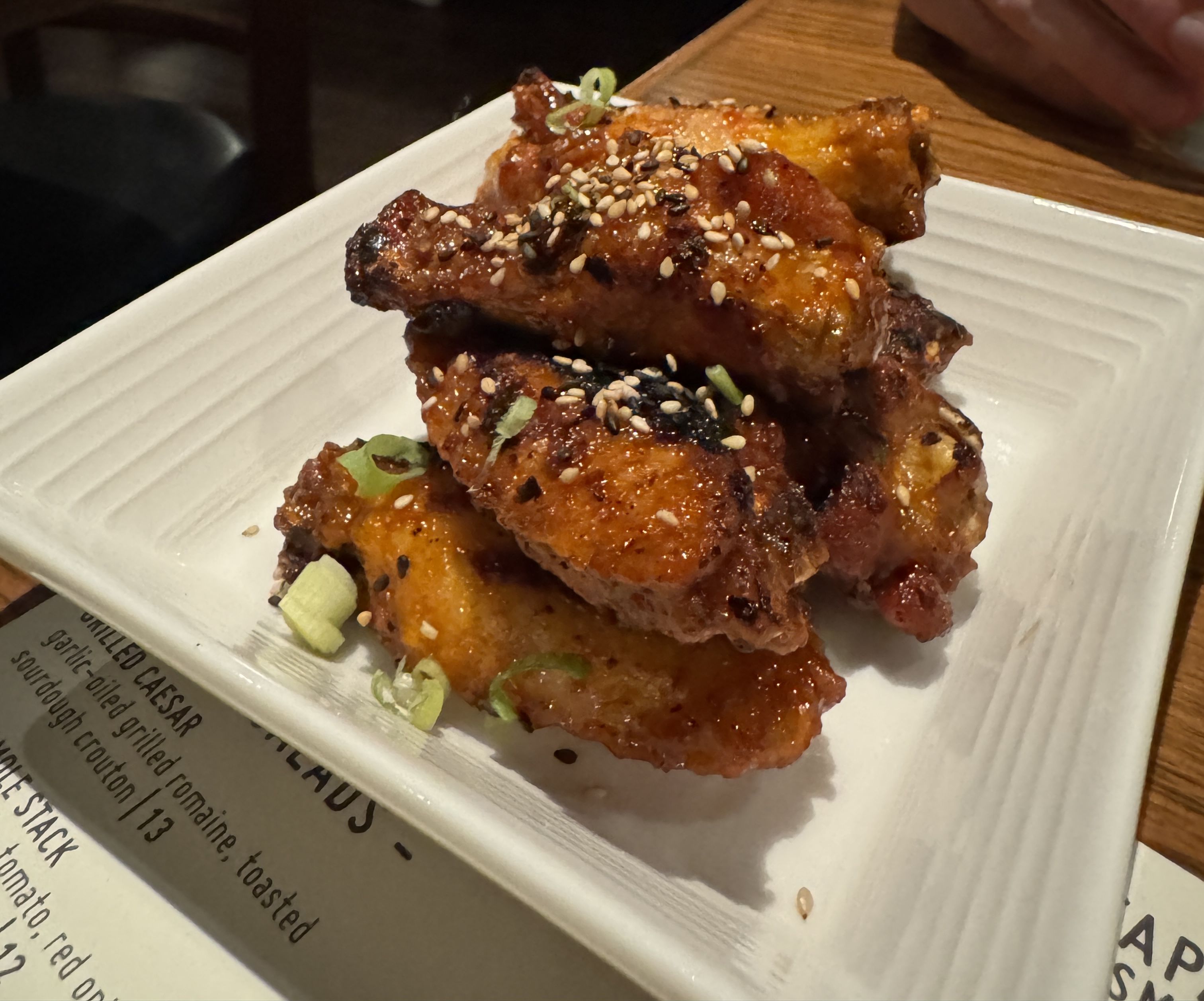 Plate of glazed chicken wings garnished with sesame seeds and chopped green onions on a white square dish, placed on a wooden table with a menu partially visible.