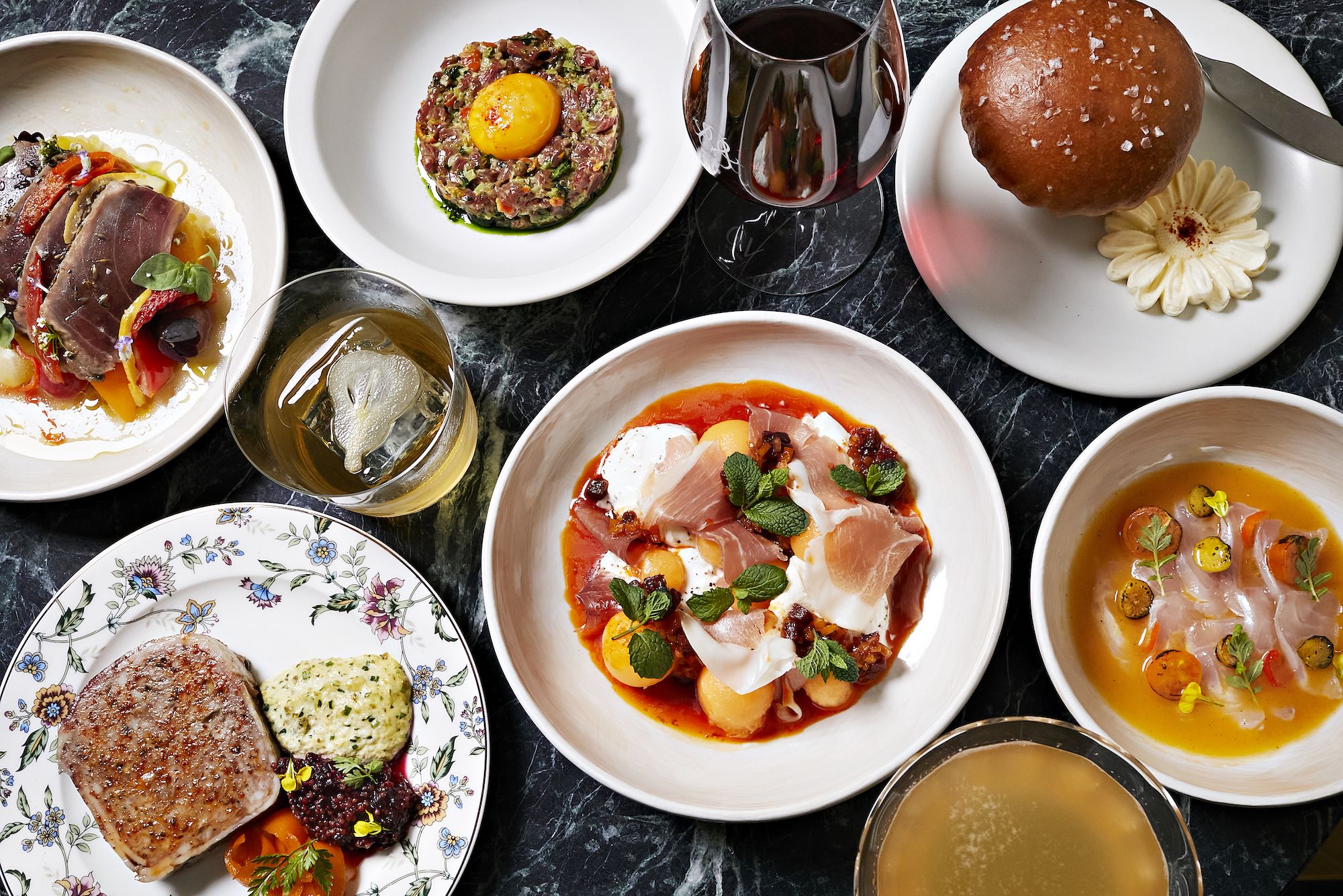 Top view of a gourmet meal spread on a black marble table including steak with sauces on floral plate, fish dish, steak tartare with egg yolk, melon and prosciutto, bread roll with flower butter, and drinks.