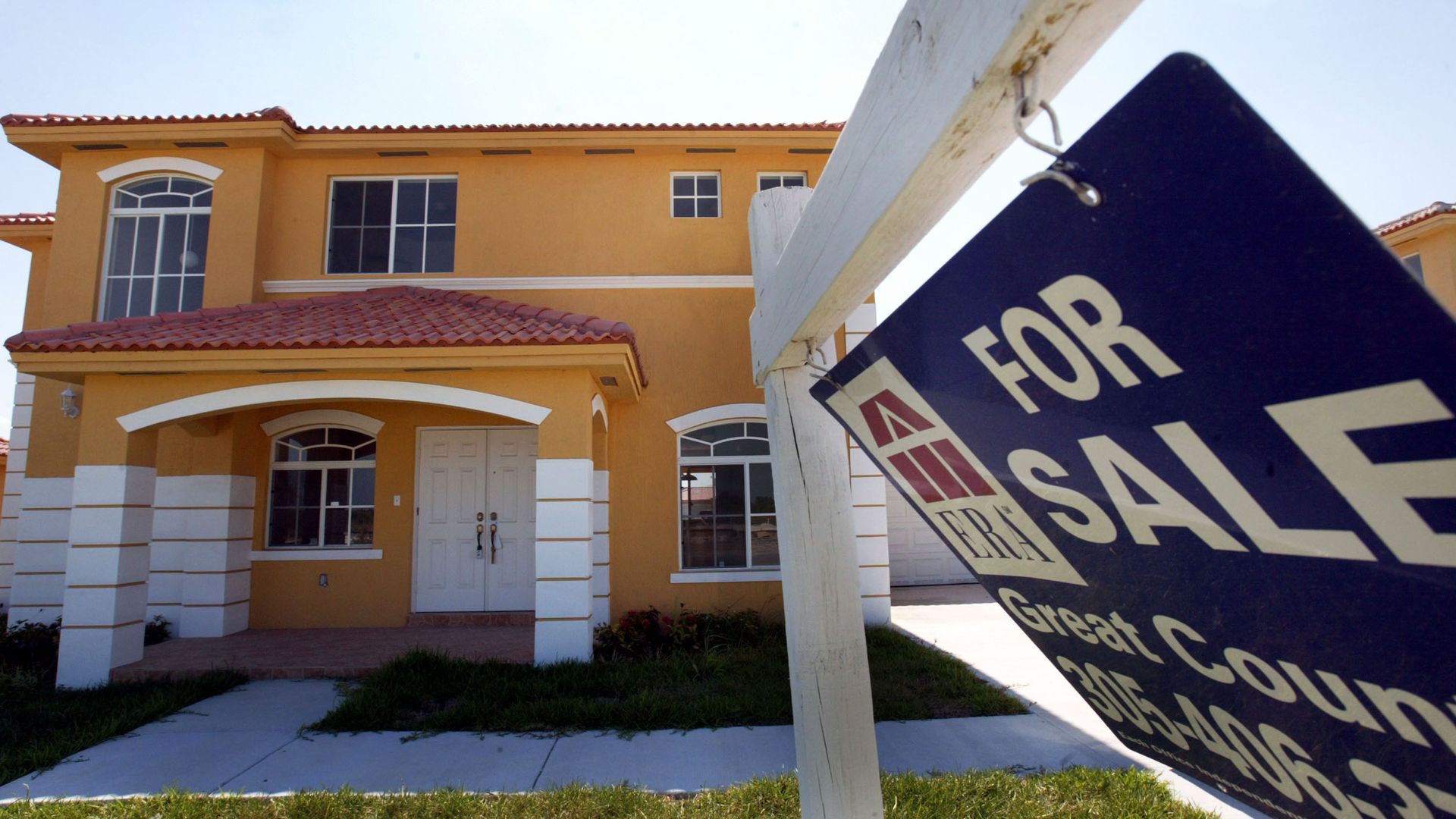 Yellow two-story house with white trim, red tile roof, and two large front windows, shown with a blue and white "For Sale" sign in the foreground on the lawn.