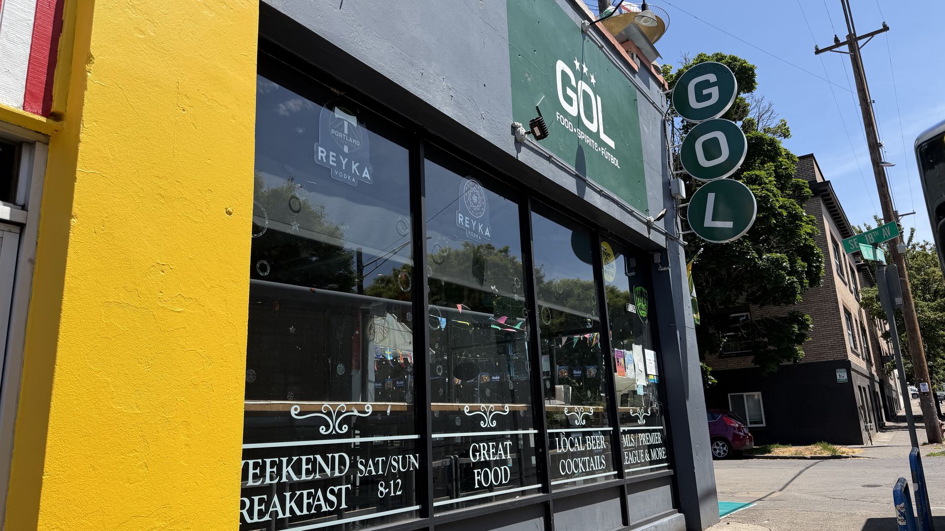 Bright storefront with a bold yellow wall on the left and a gray-green facade. Windows advertise weekend breakfast and food; three green circular signs hang on a pole under a blue sky.