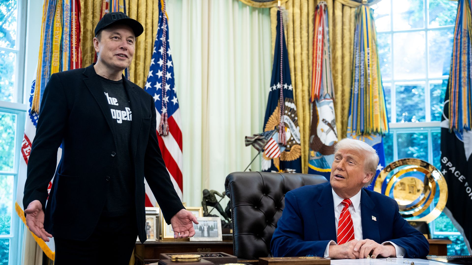 Elon Musk stands next to President Trump, seated at desk