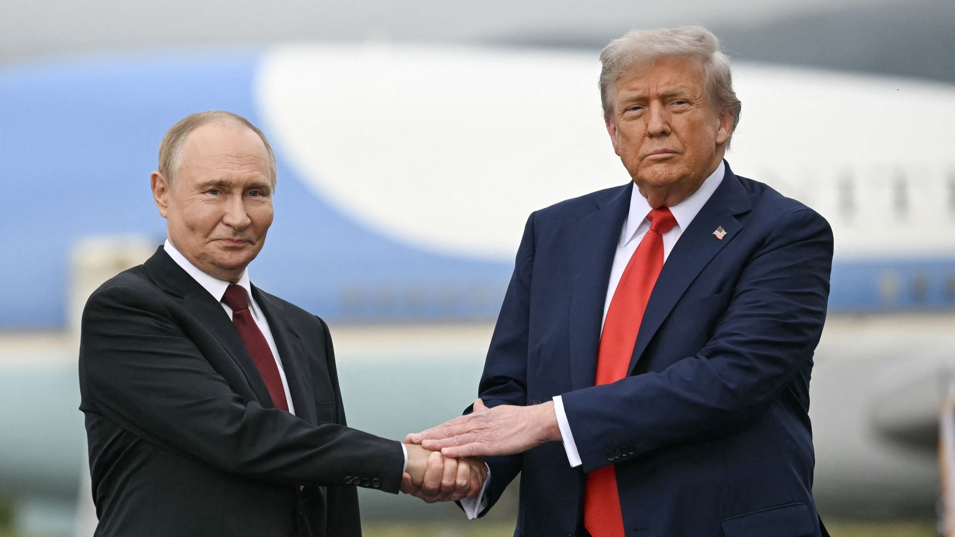 Trump, wearing a dark suit and a red tie, places his hand over Putin's as the two men shake hands.