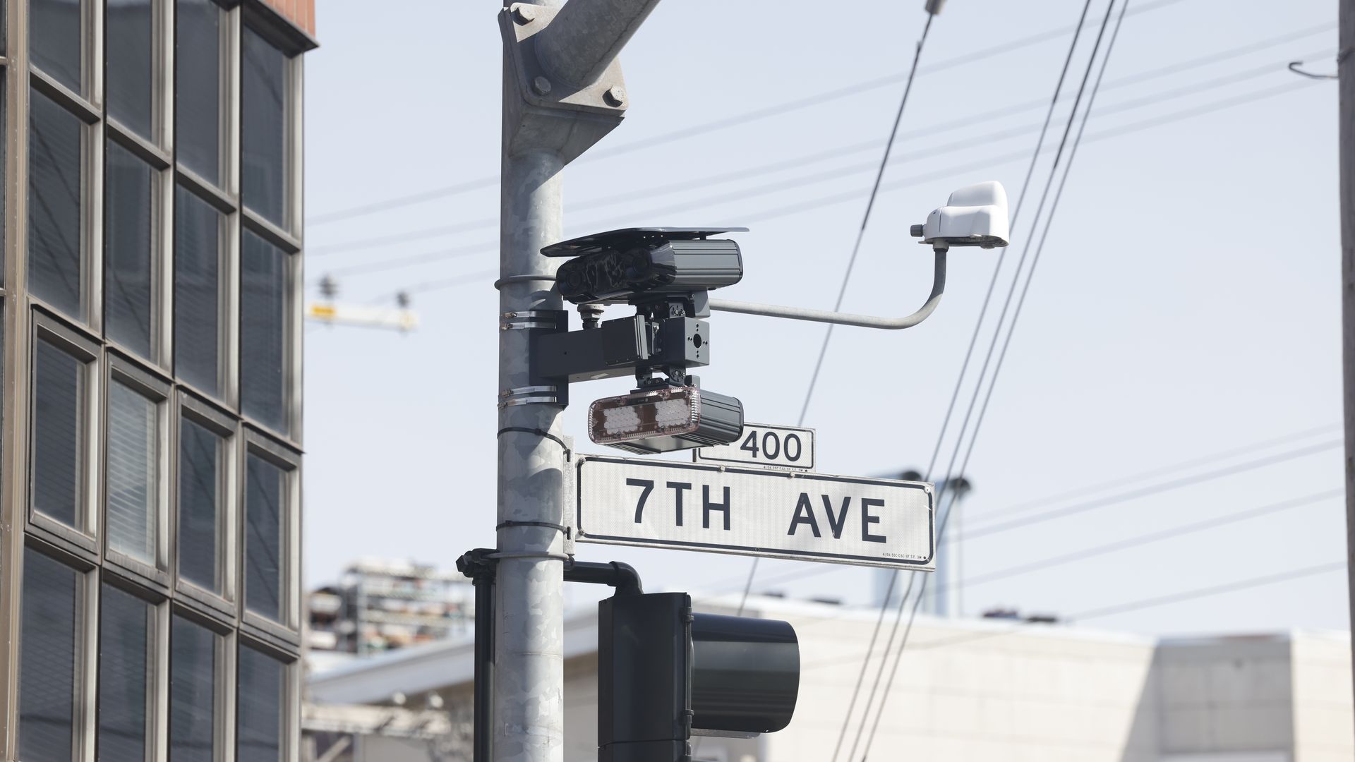 Traffic camera and 7TH AVE street sign on a metal pole near a building with glass windows, under clear sky with visible power lines in urban setting.