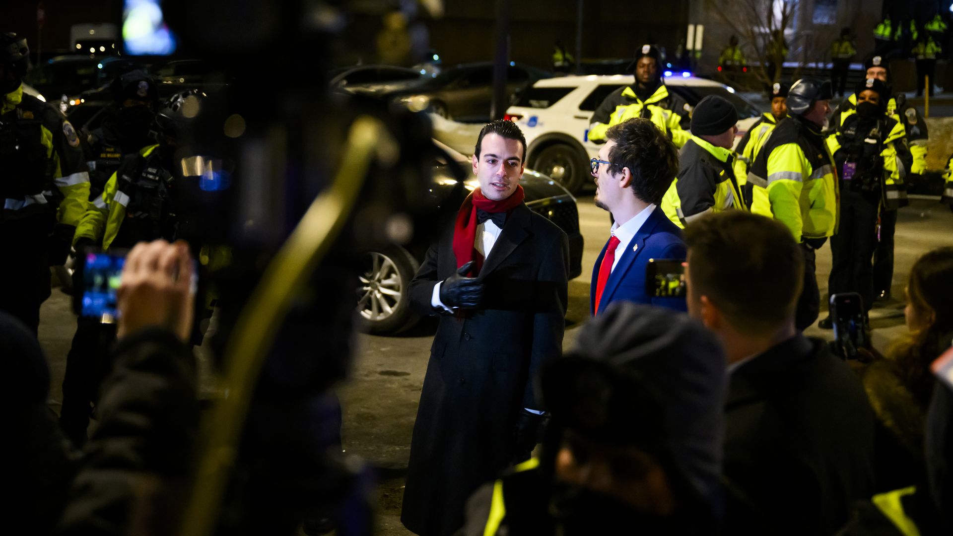 Police officers and reporters stand near two men on a city street. One wears a black jacket, black gloves and a red scarf, while the other wears a blue suit, white shirt and red tie.