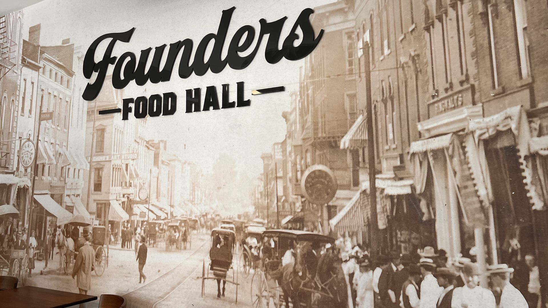 Sepia-toned mural of a vintage city street with storefronts, pedestrians, and horse-drawn carriages. A large black cursive sign reads "Founders" with "FOOD HALL" beneath; foreground shows cafe tables.