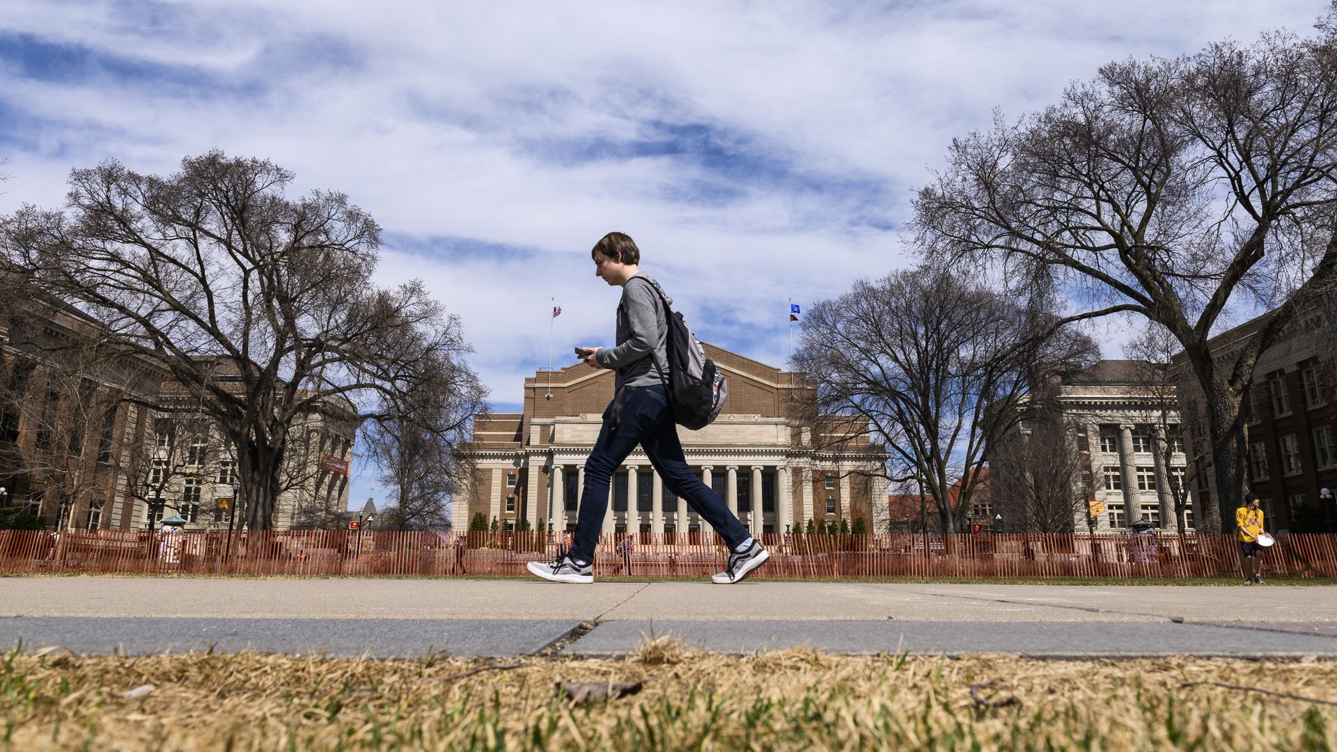 A student in a gray long-sleeved shirt and sneakers walks across a college campus with a red-brown brick building and columns in the background.