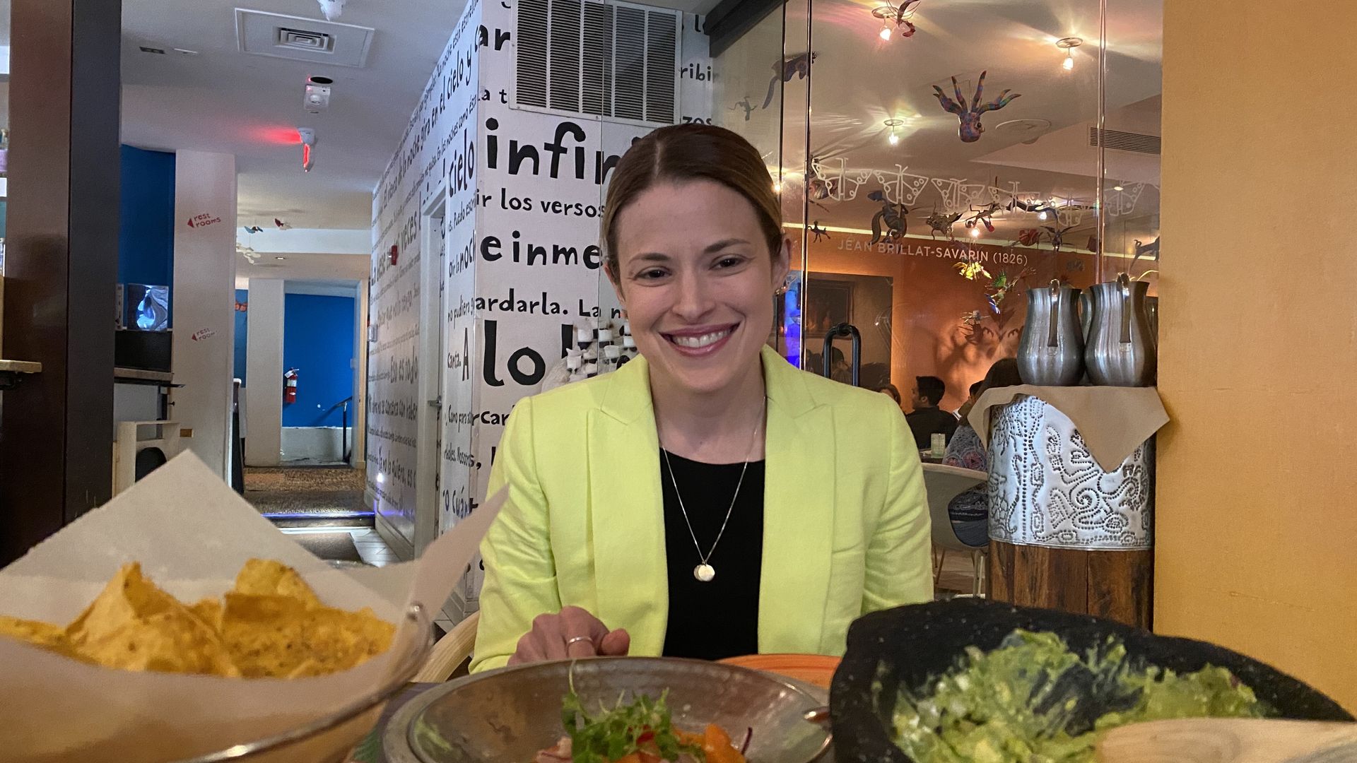 Niki Carelli, a white woman in a bright green blazer over a black shirt, sits in front of a spread of Mexican food.