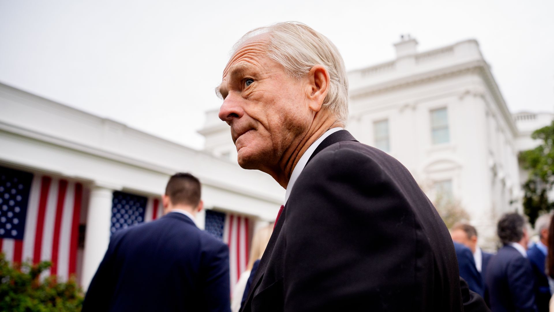 Man in a dark suit standing in front of the White House, draped in U.S. flags.