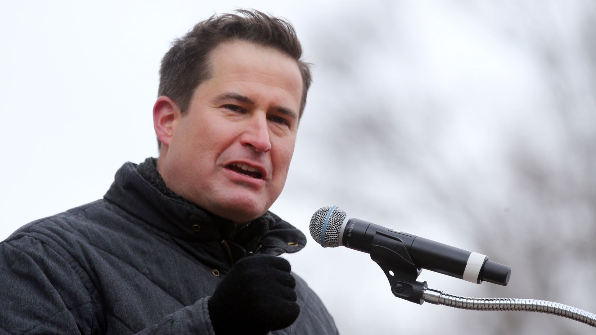 In this image, Seth stands in a winter coat outside and speaks into a microphone while gesturing with one hand.