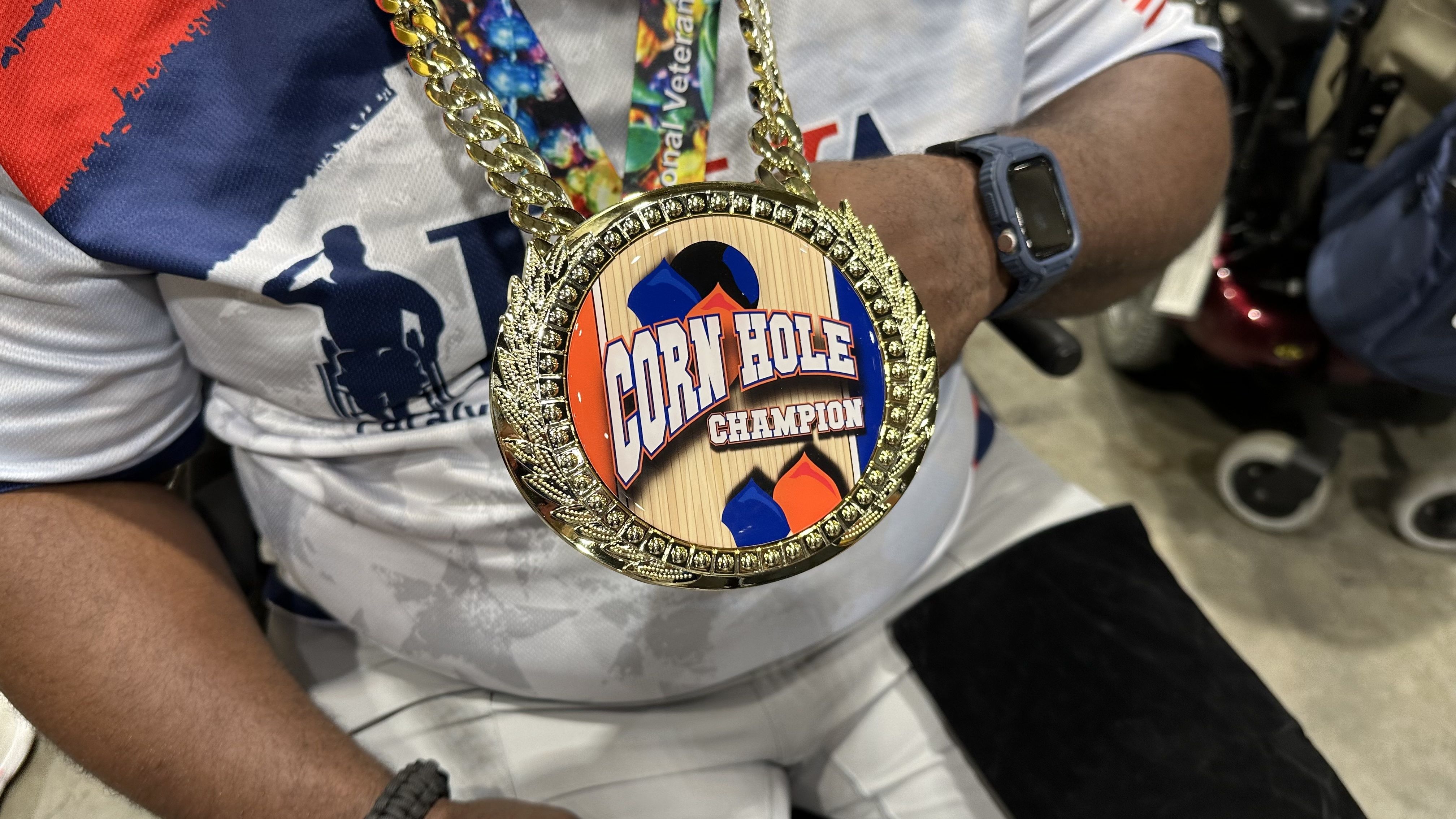 A man holds up a large gold medal that says "Corn Hole Champion."