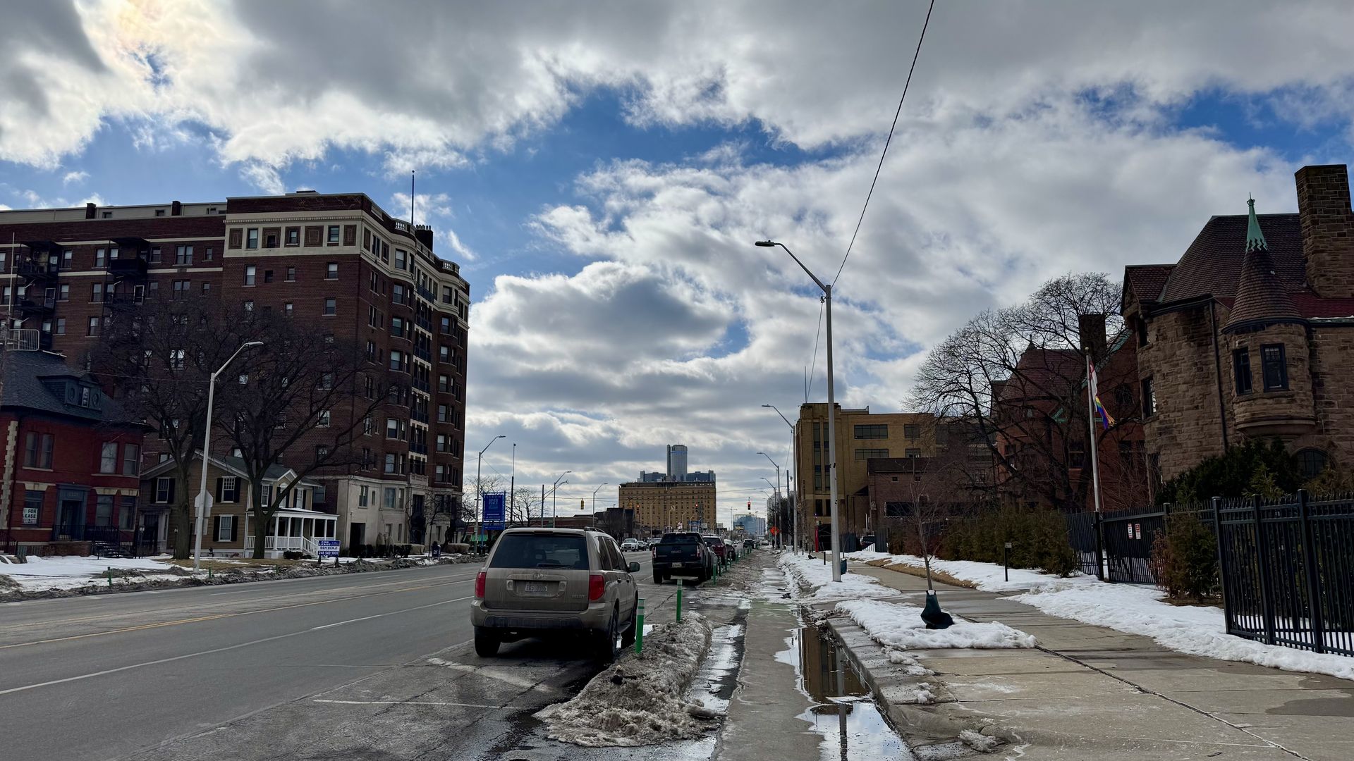 Urban street with parked cars, wet snow piles, and puddles. Brownstone buildings line the street under a partly cloudy blue sky with patches of sunlight.